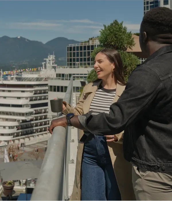A woman holding a mug and smiling chats with a man on a rooftop balcony, with a cityscape, cruise ship, and mountains in the background—perfect inspiration for your Vancouver itinerary.