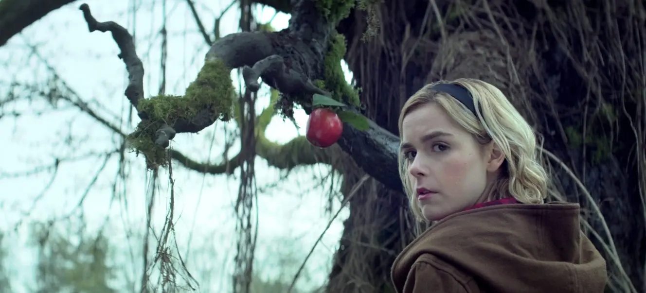 Person with blonde hair and brown coat looking at a red apple hanging from a moss-covered tree branch.