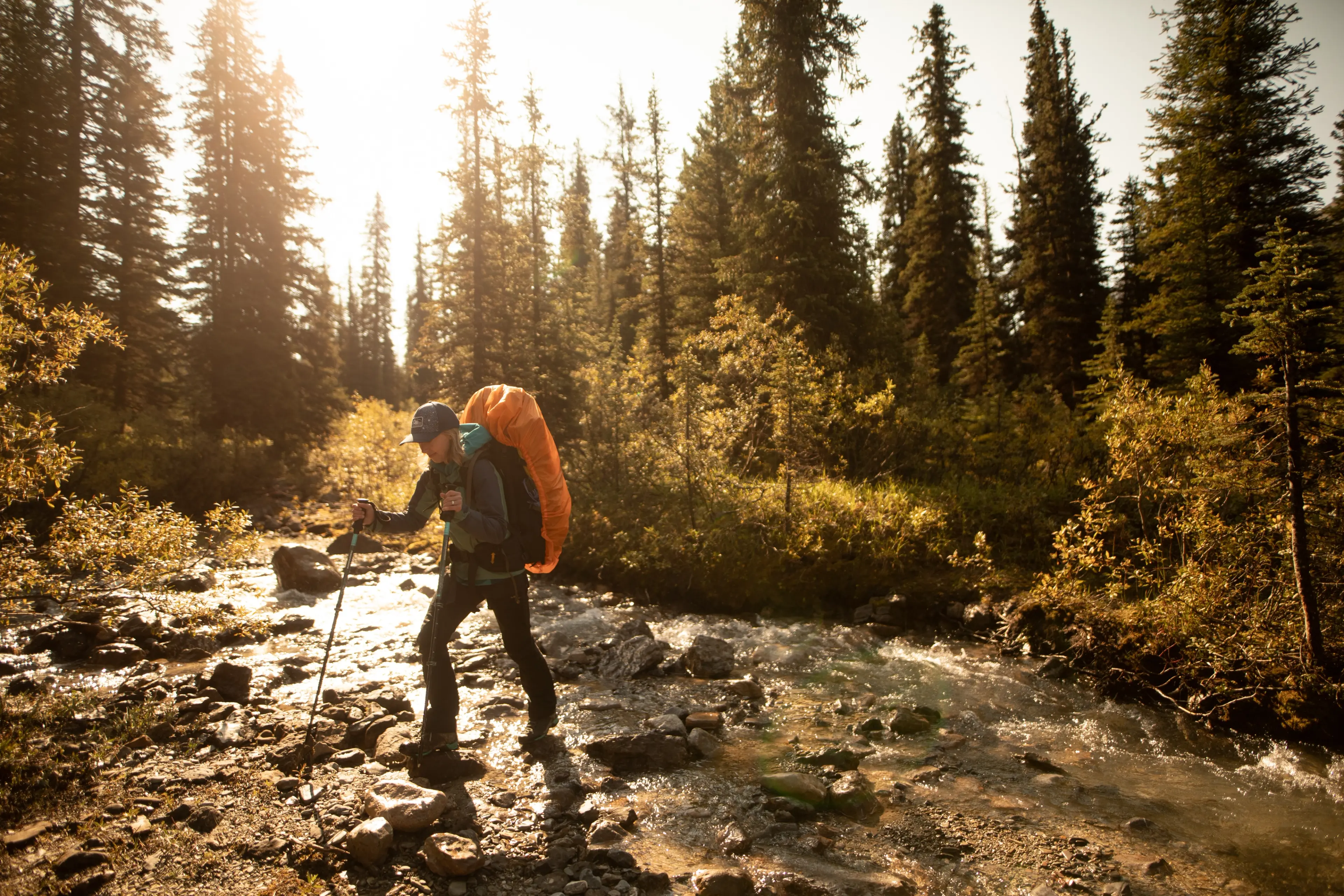 A woman wearing a backpack crosses a stream in a forest in a still from the film Wildflowers screening at the 2025 Vancouver International Mountain Film Festival