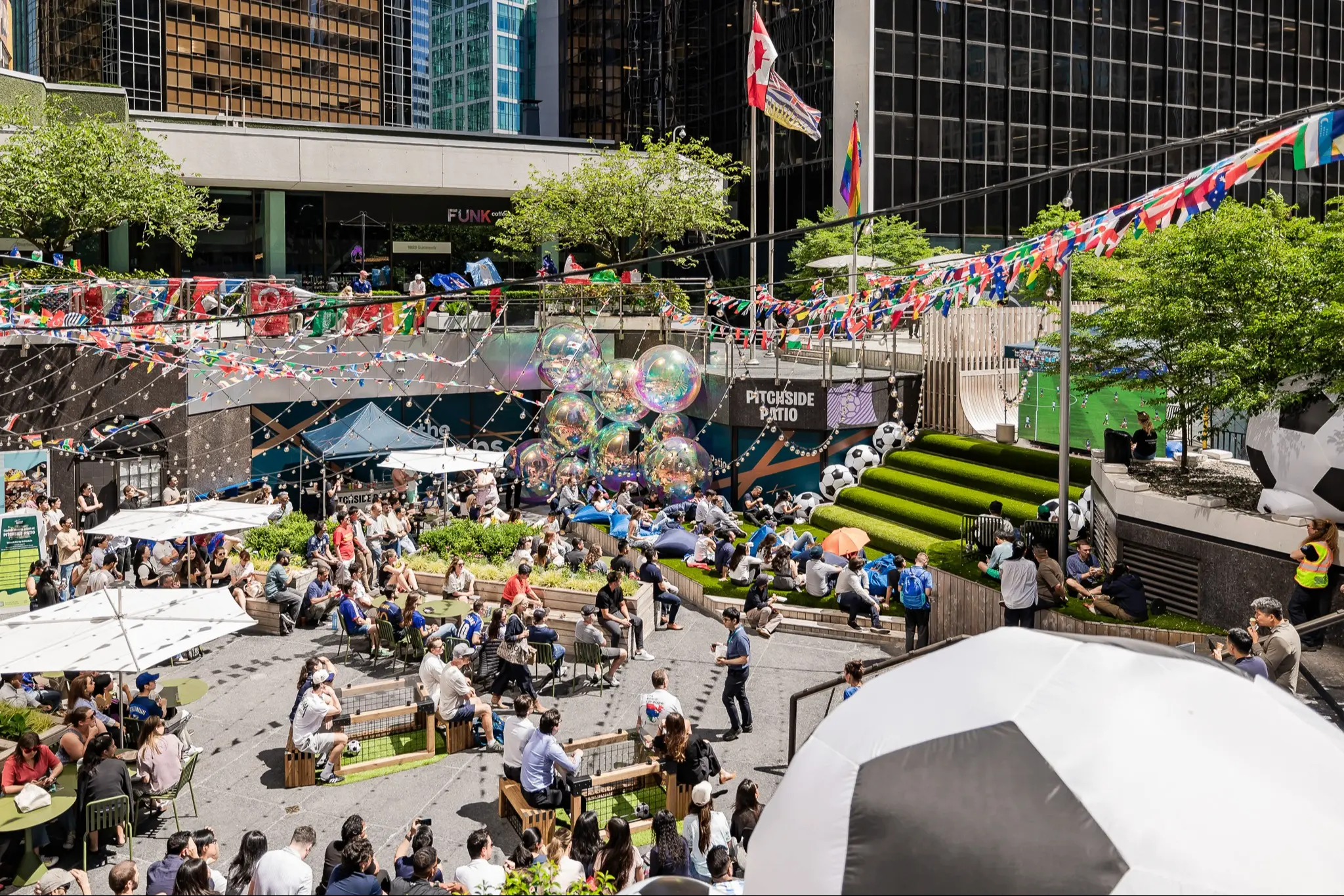 Outdoor patio with many people seated under umbrellas and colorful flags, decorated with large soccer balls and shiny balloons.