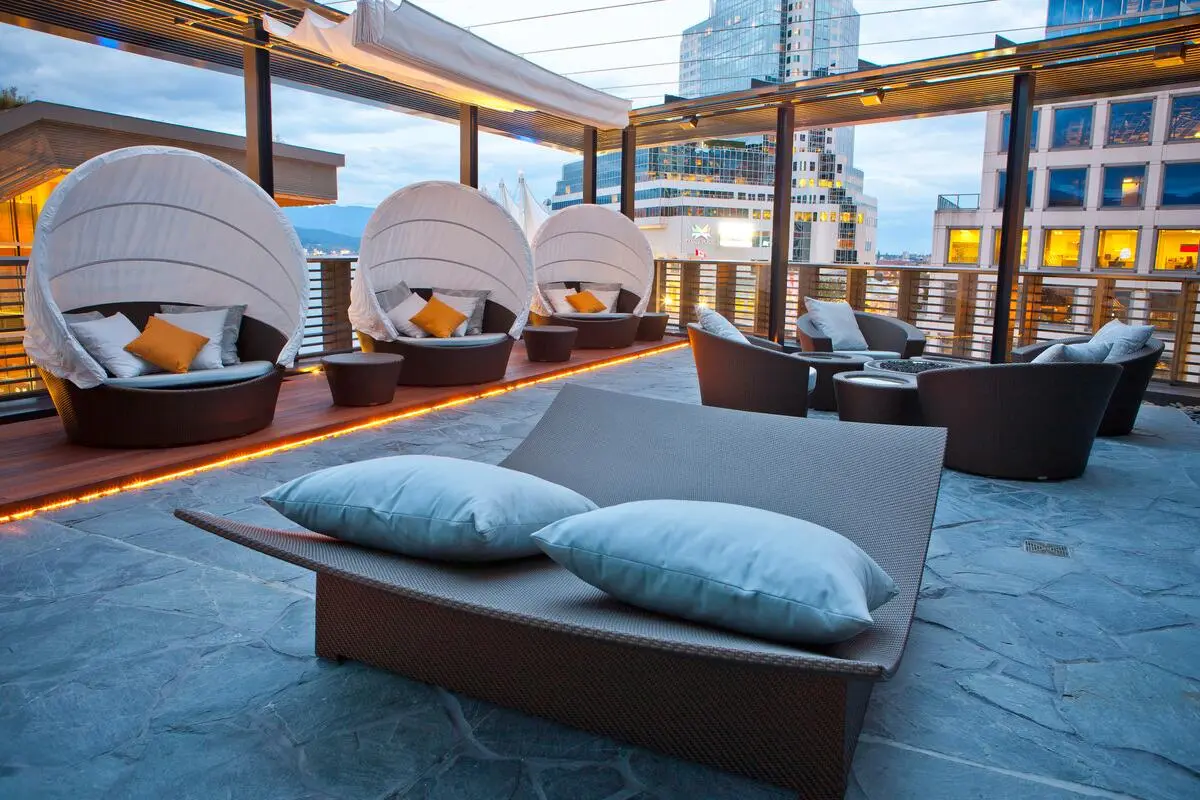 Modern rooftop lounge with curved wicker chairs, white cushions, and city buildings in the background at dusk.