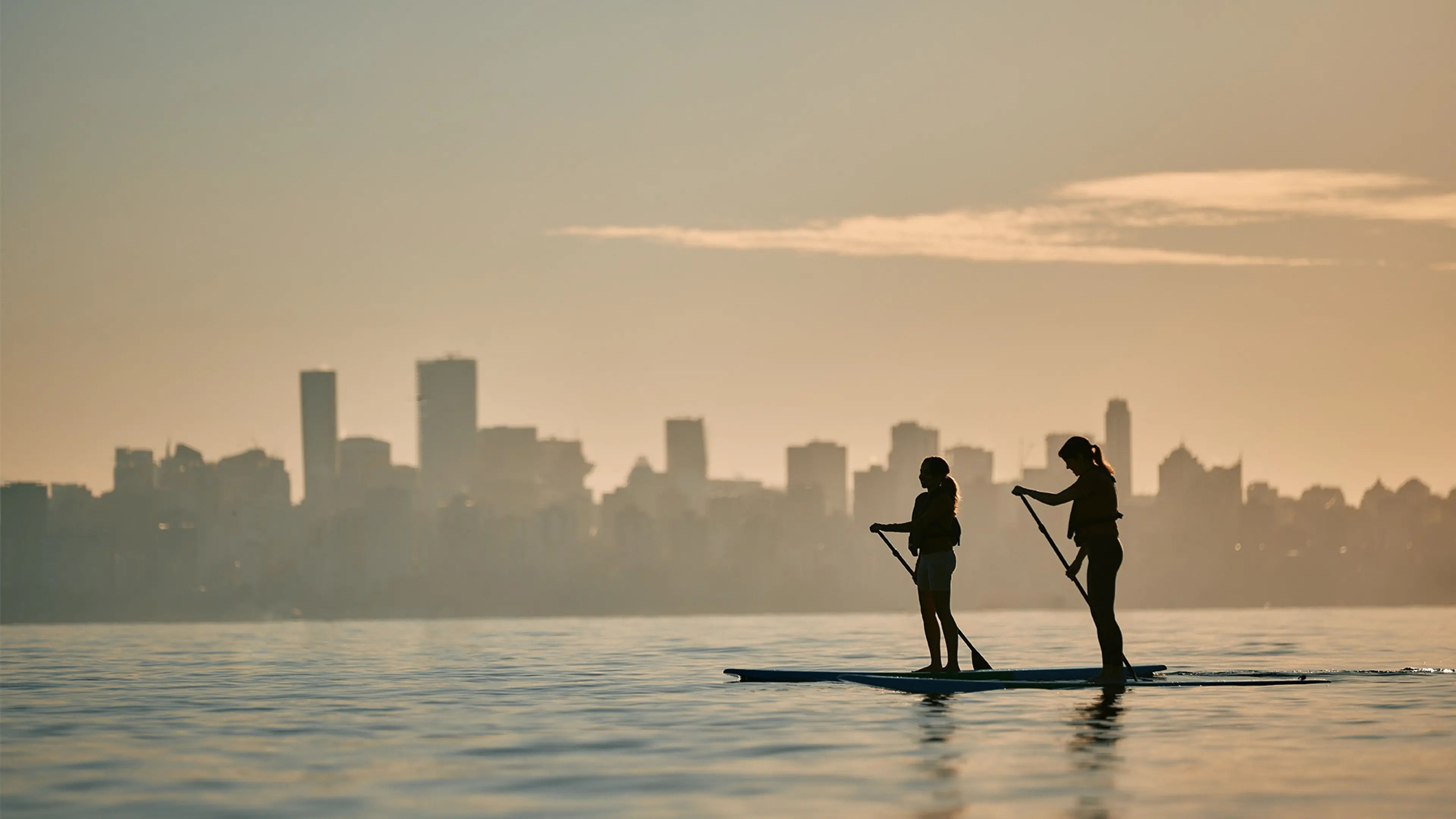 Two people paddleboarding on calm water with a city skyline in the background at sunset.