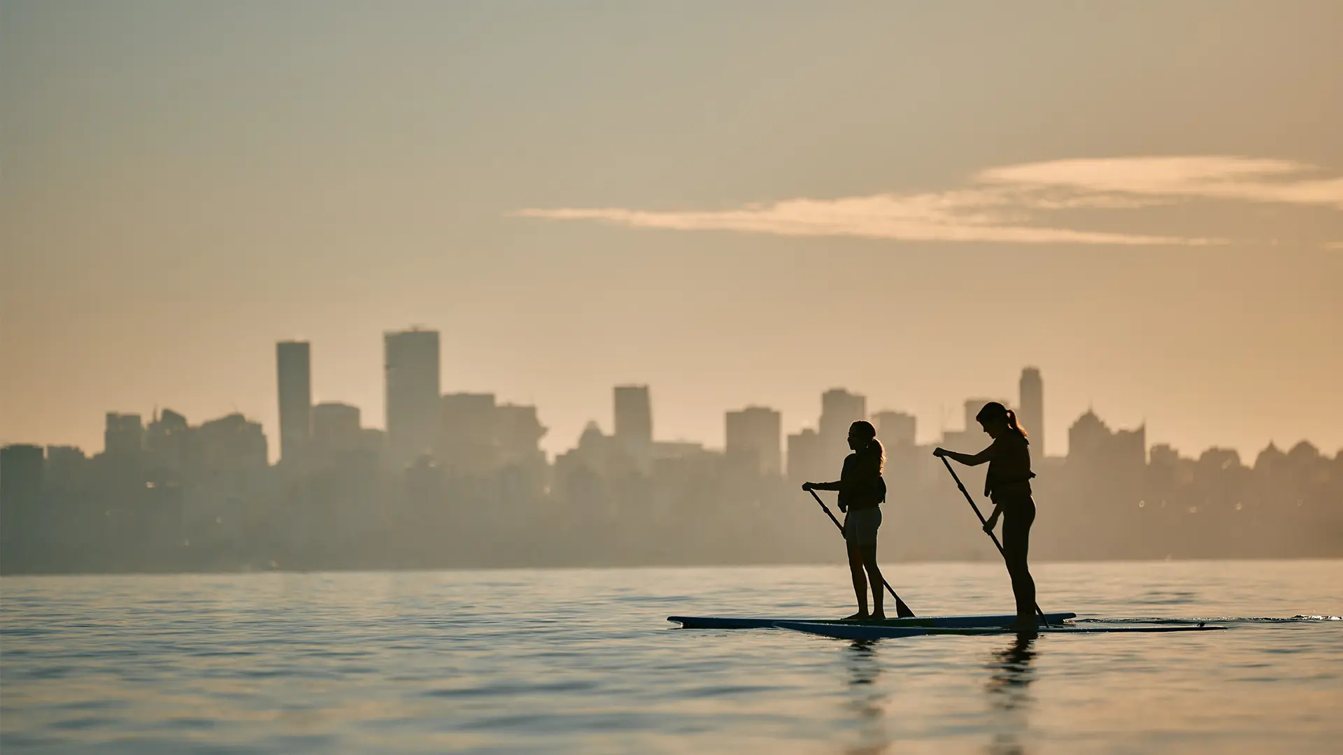 Two people paddleboarding on calm water with a city skyline in the background at sunset.