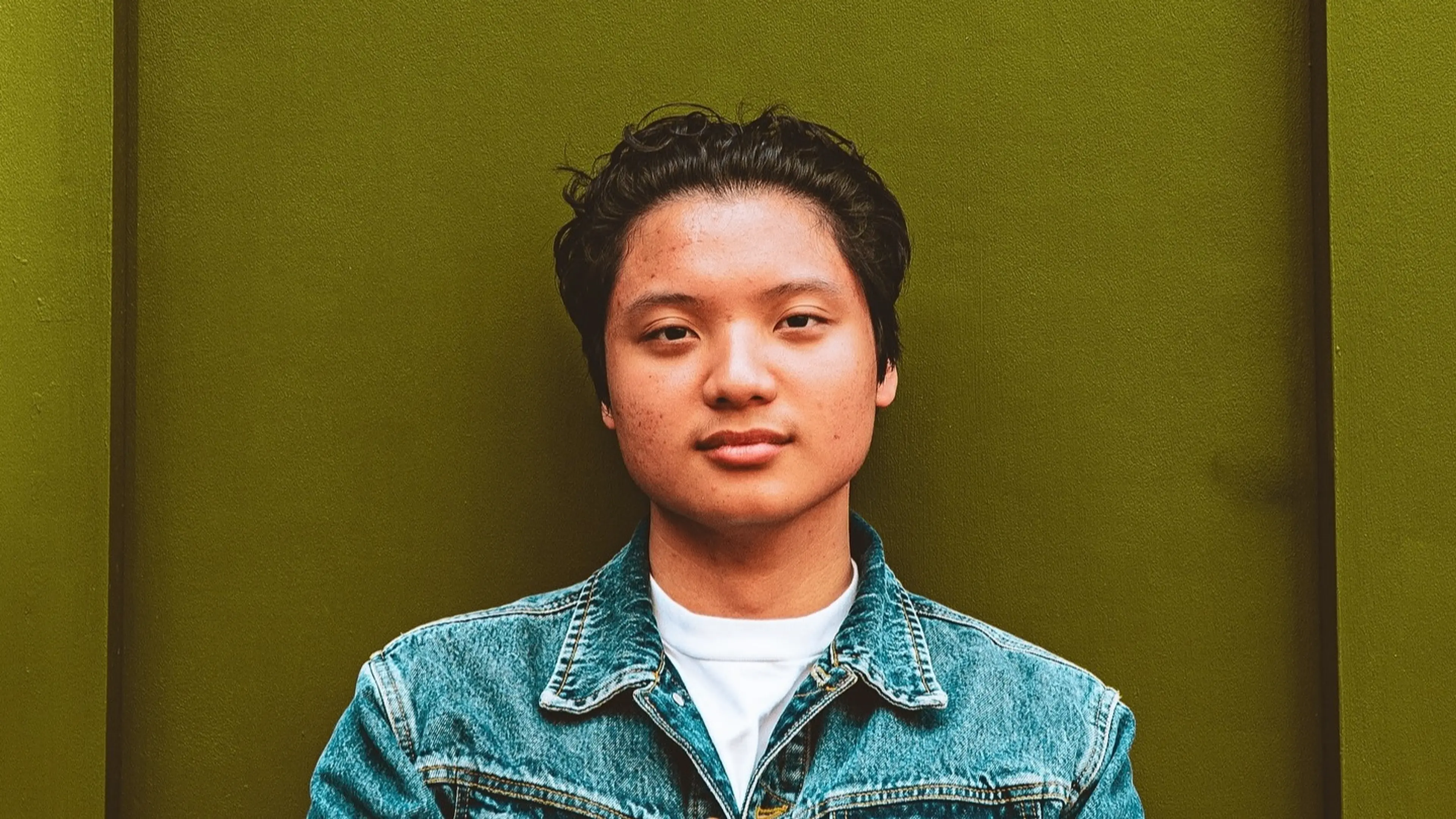 Person with short curly hair wearing a blue denim jacket and white shirt against an olive green background.
