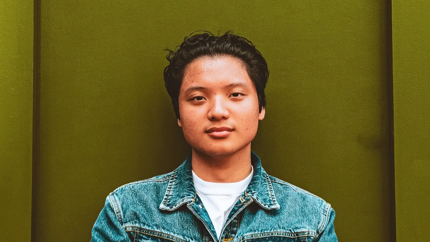 Person with short curly hair wearing a blue denim jacket and white shirt against an olive green background.