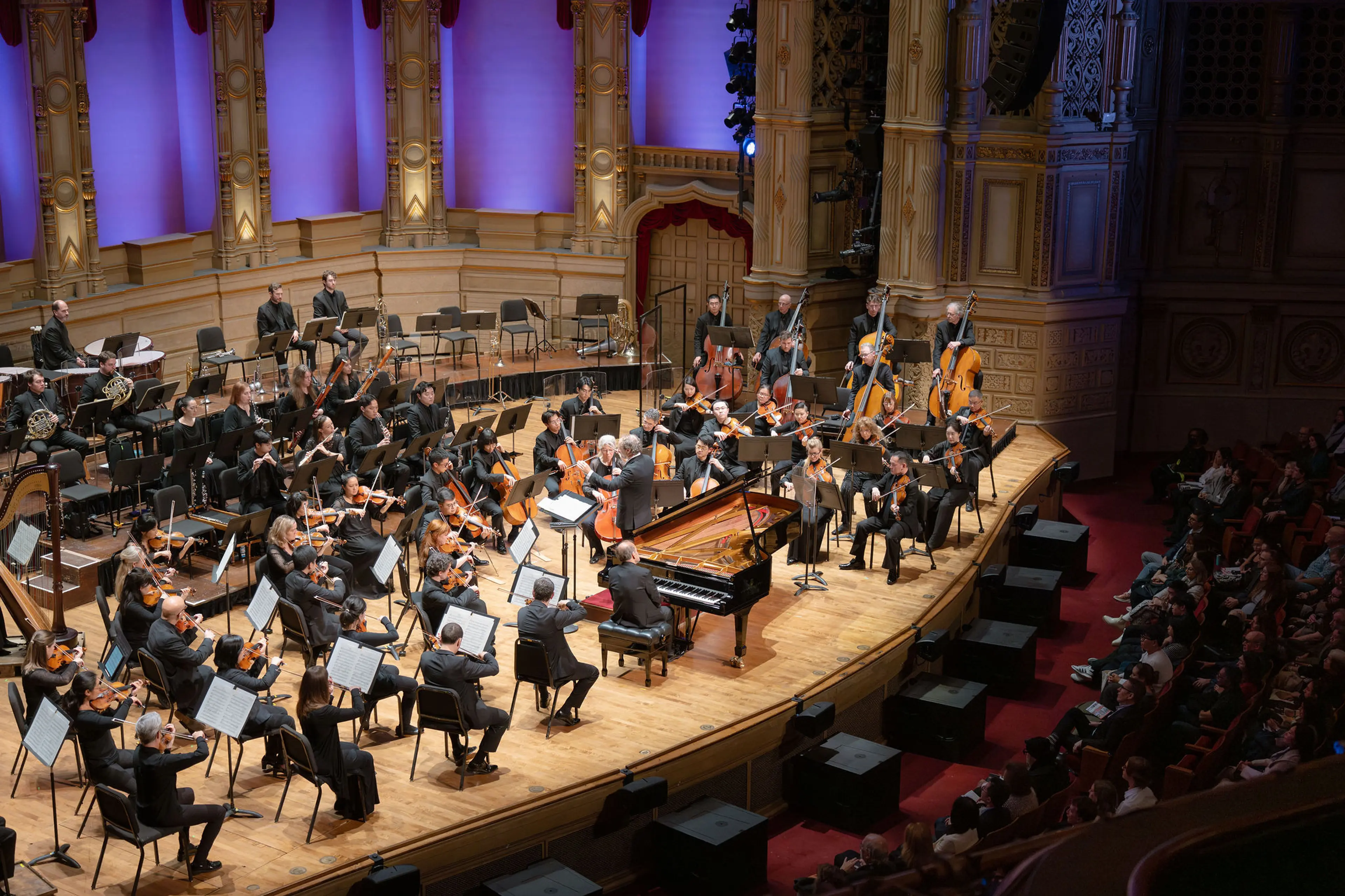 Orchestra performing on stage with conductor and pianist in a grand concert hall with audience seated.