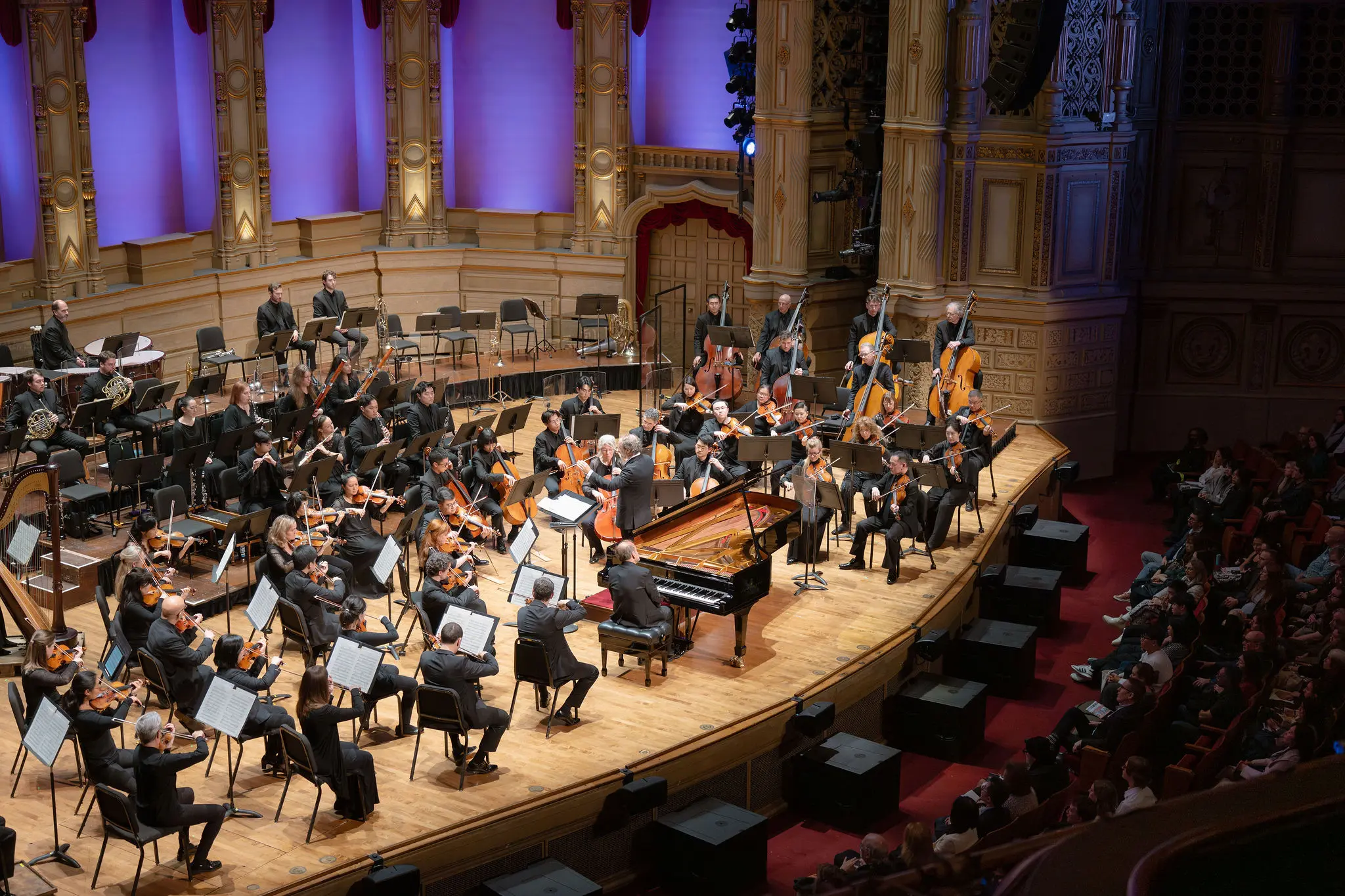 Orchestra performing on stage with conductor and pianist in a grand concert hall with audience seated.