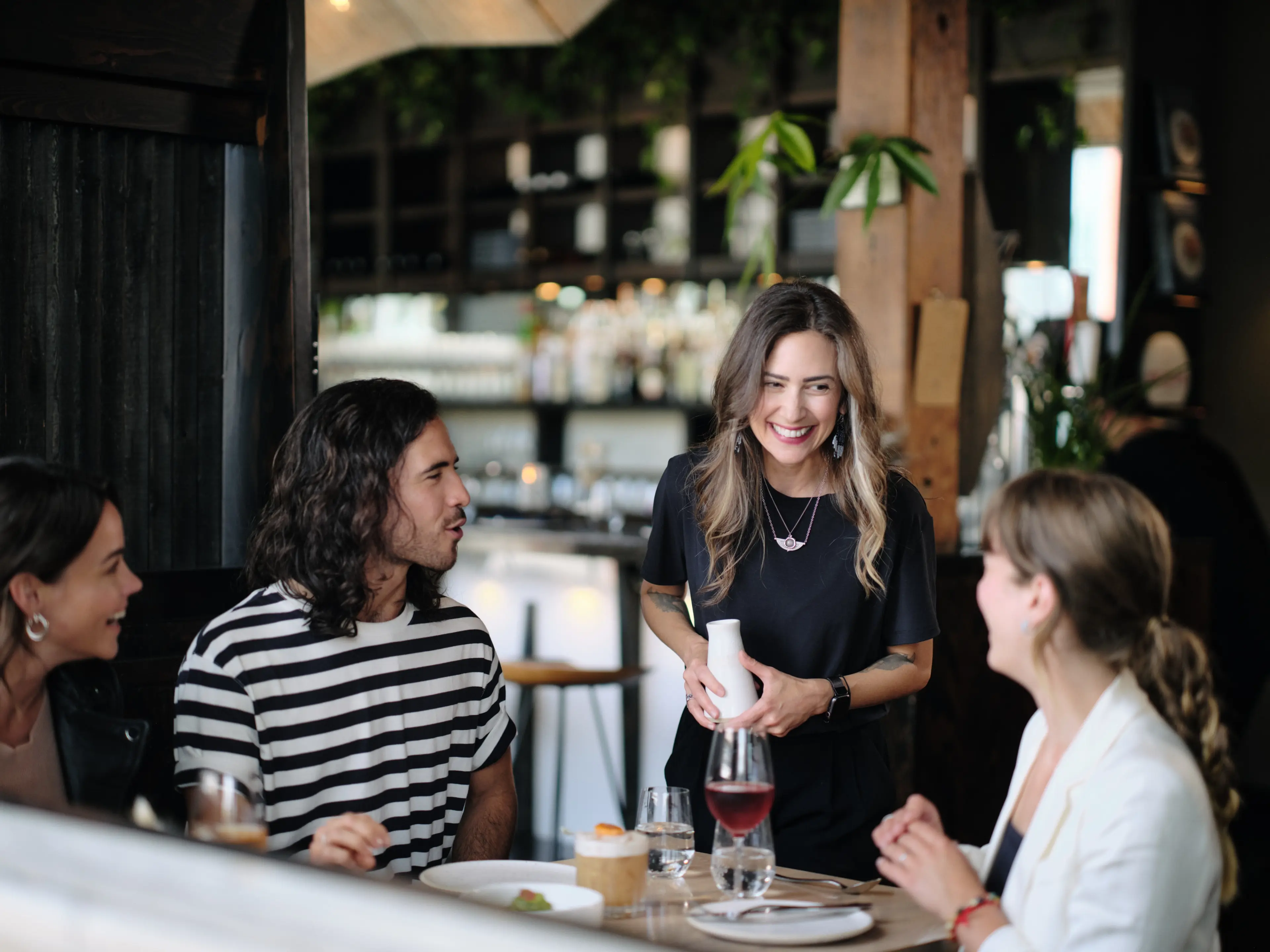 Four people at a restaurant table, one standing and smiling while holding a white bottle, others seated and engaged in conversation.