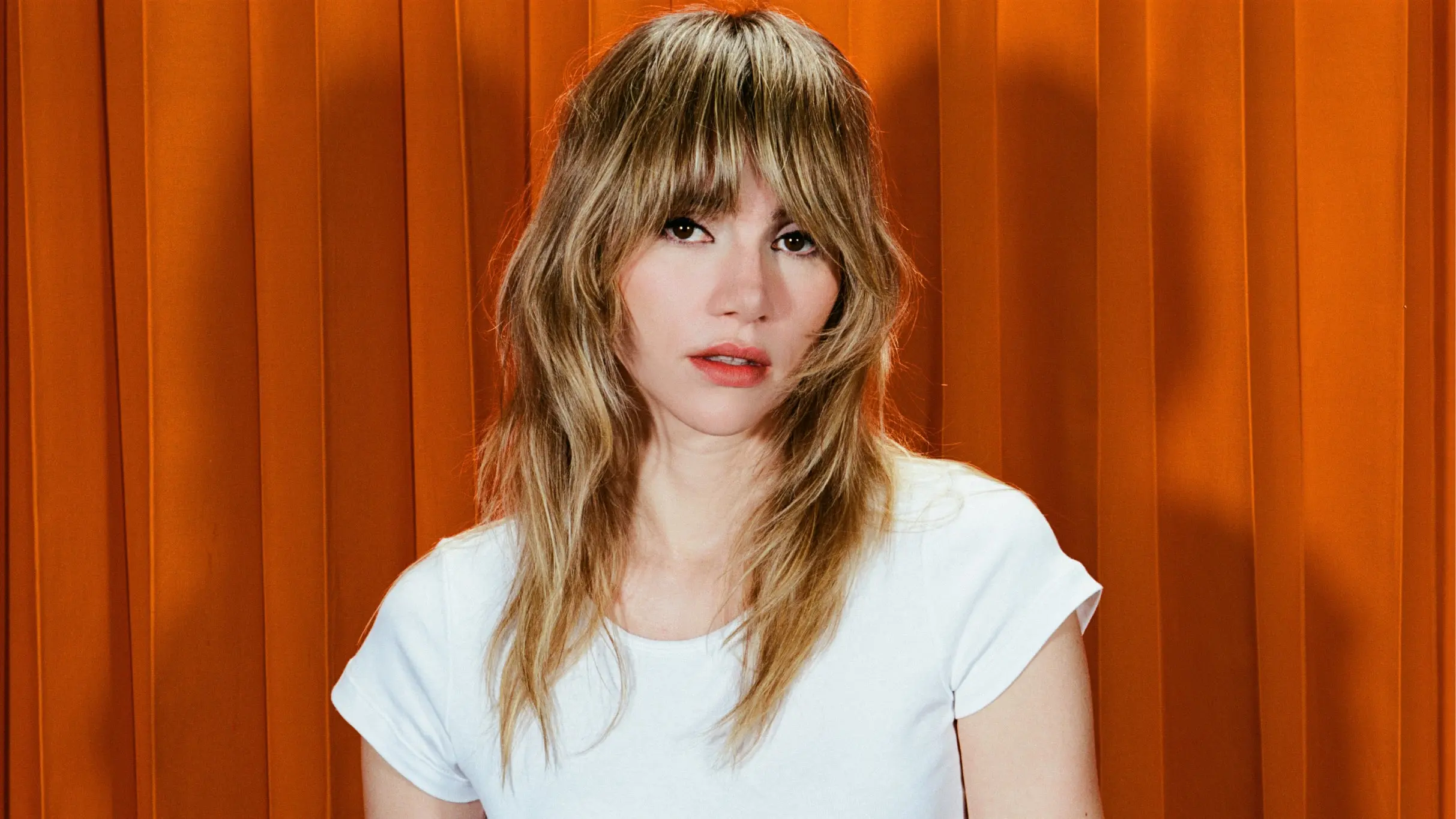 Person with long, wavy light brown hair wearing a white t-shirt against an orange curtain background.