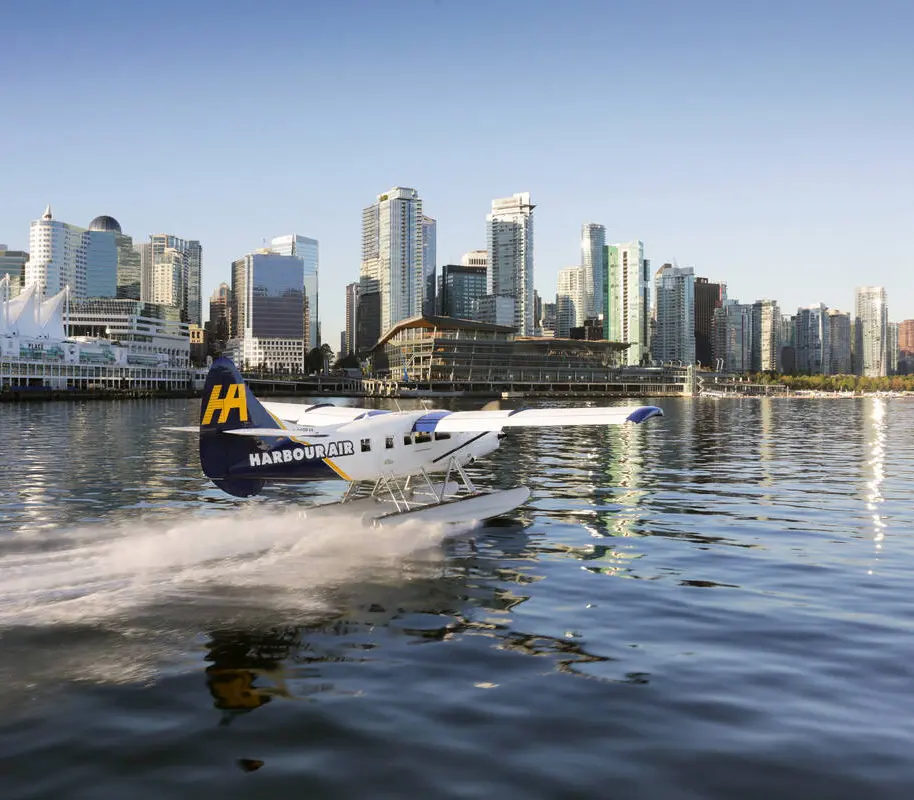 A seaplane with Harbour Air branding flies over calm blue water, passing rocky, tree-covered islands—a perfect scene to inspire your trip to Vancouver under a cloudy sky.