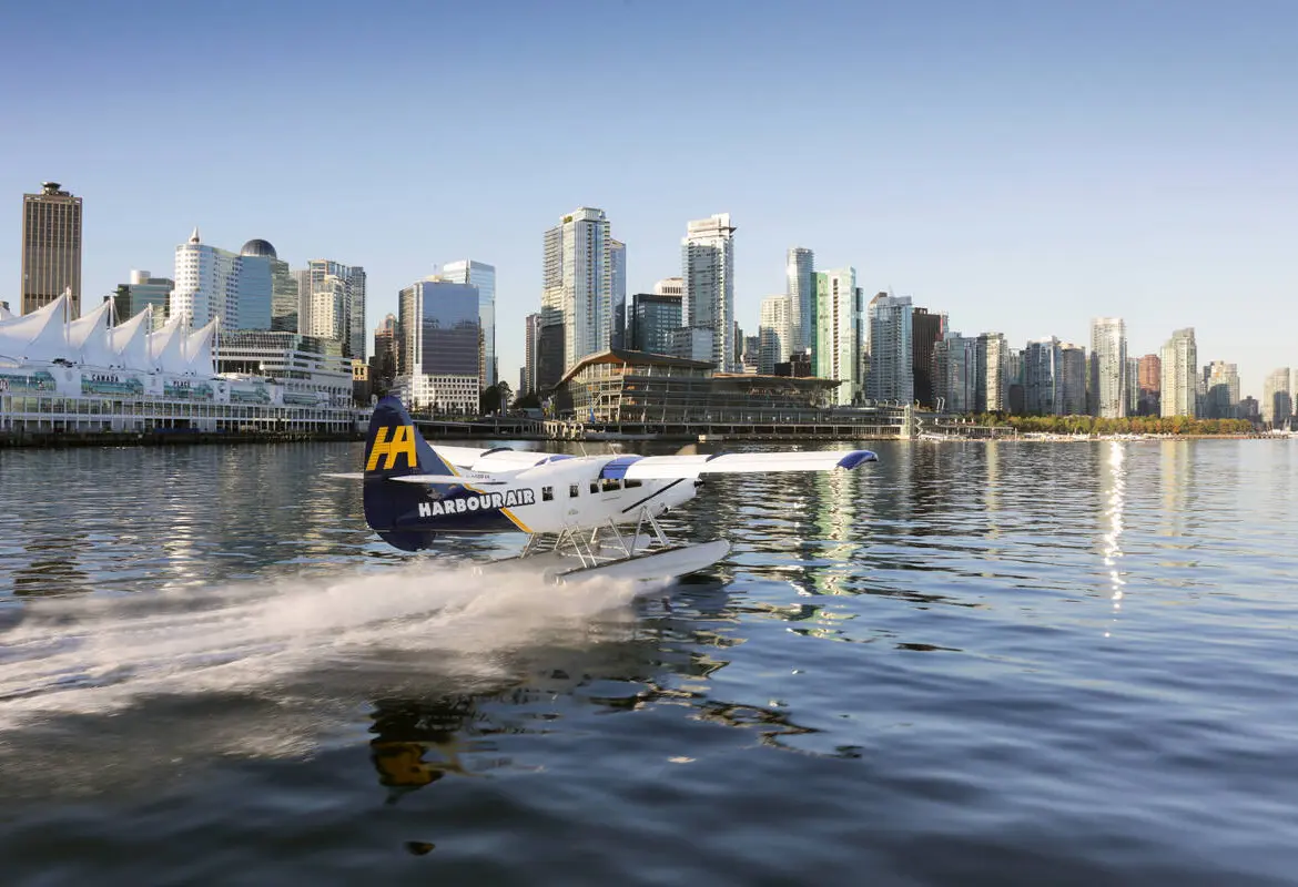 A seaplane with Harbour Air branding flies over calm blue water, passing rocky, tree-covered islands—a perfect scene to inspire your trip to Vancouver under a cloudy sky.
