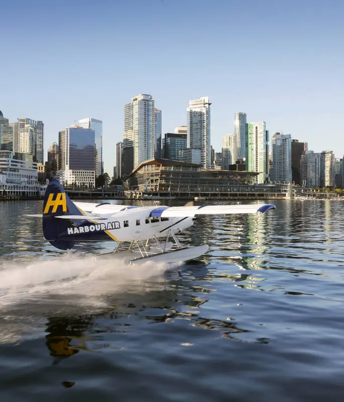 A seaplane with Harbour Air branding flies over calm blue water, passing rocky, tree-covered islands—a perfect scene to inspire your trip to Vancouver under a cloudy sky.