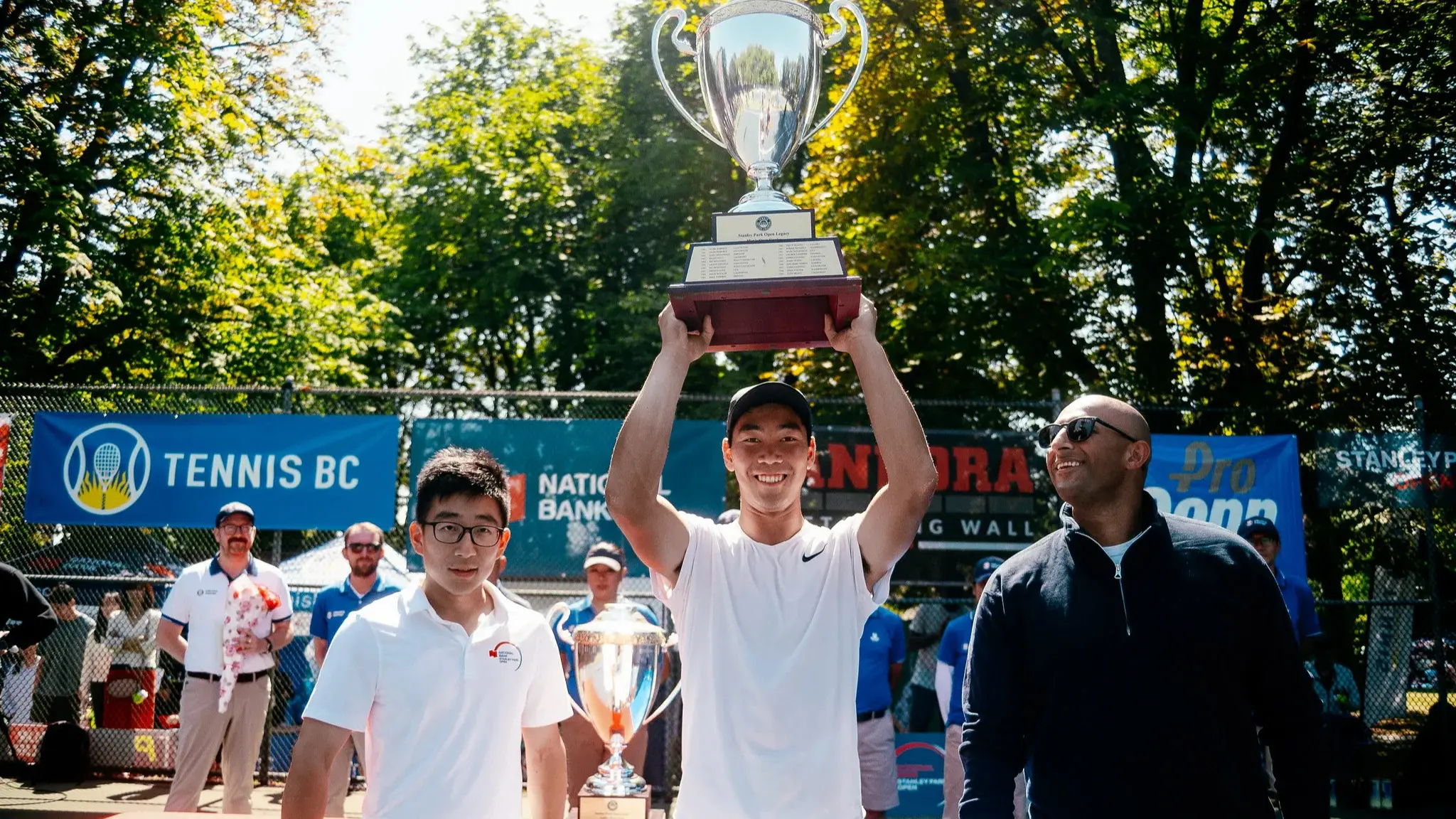 Three people standing outdoors with one person in the center holding a large trophy above their head, smiling, with tennis banners in the background.