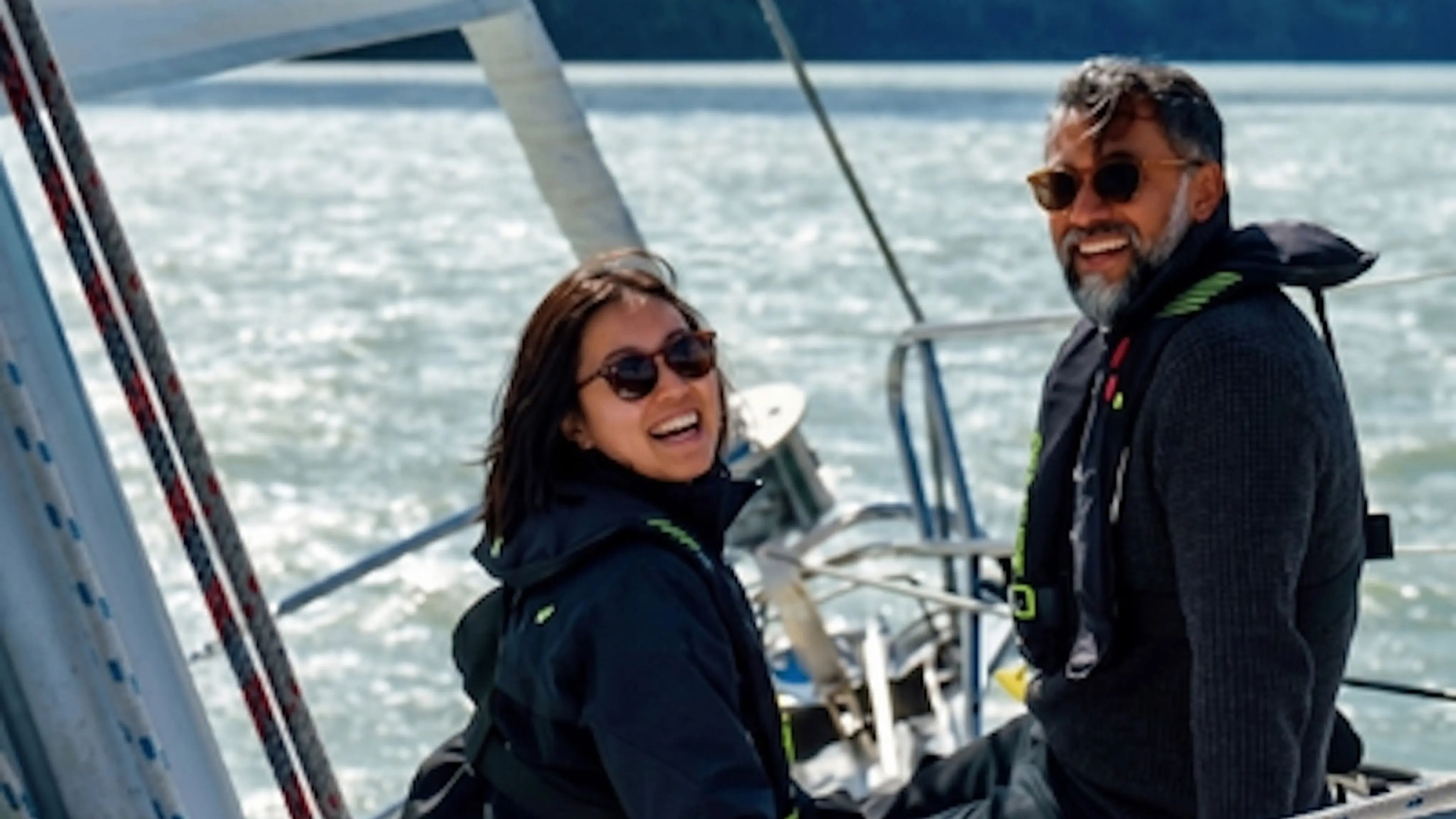 Two people wearing sunglasses and dark jackets smiling on a sailboat with water in the background.