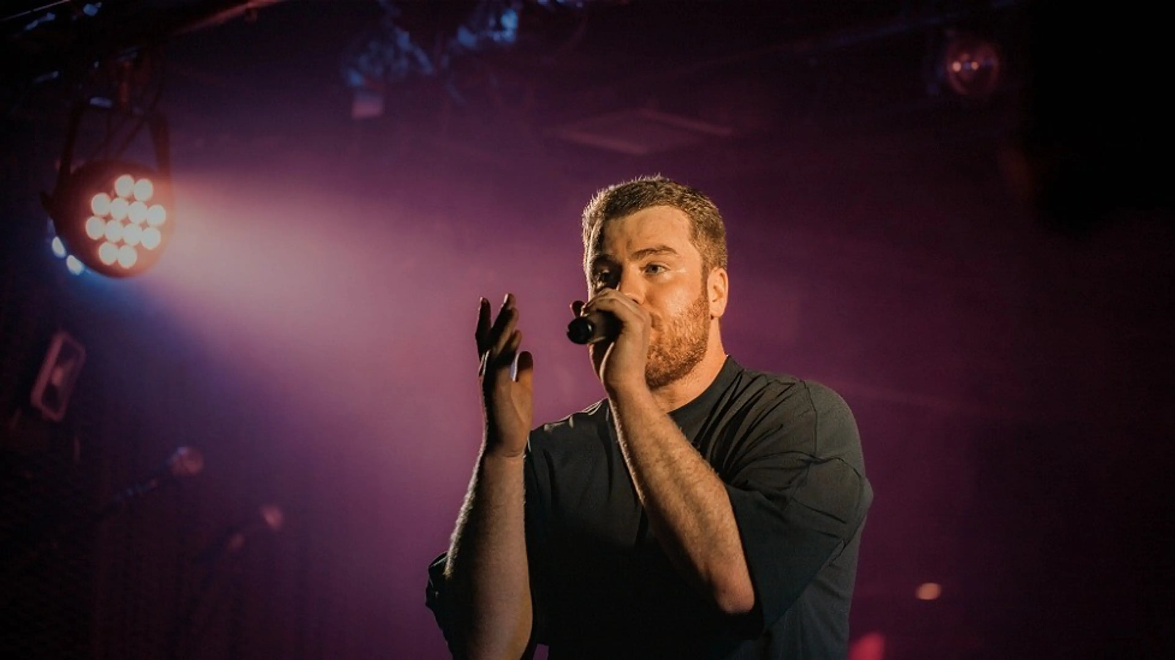 A person with short hair and beard singing into a microphone on stage with purple lighting and spotlight.