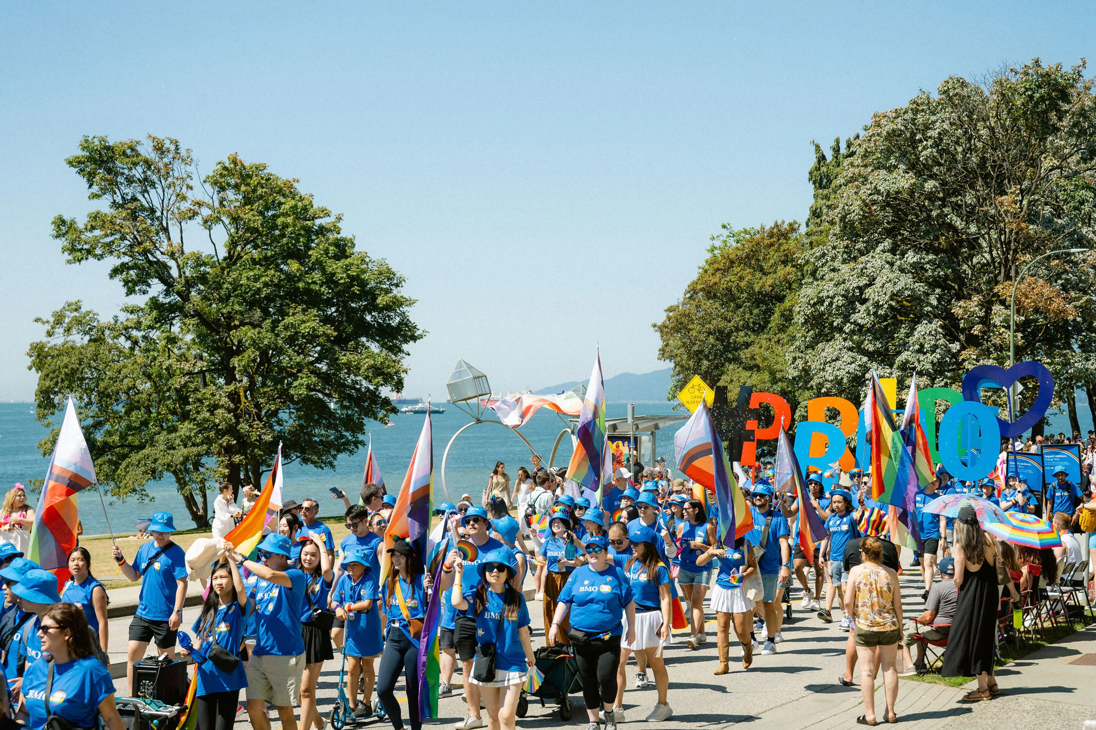 Large group of people in blue shirts marching with colorful flags and large letters spelling PRIDE near a waterfront park.