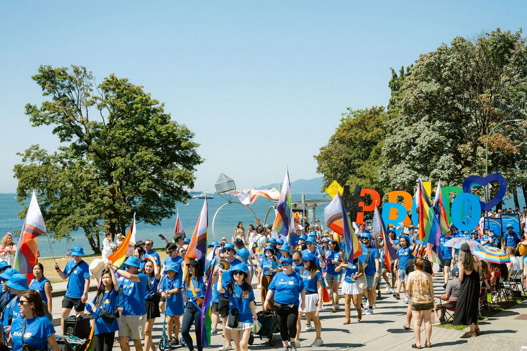Large group of people in blue shirts marching with colorful flags and large letters spelling PRIDE near a waterfront park.