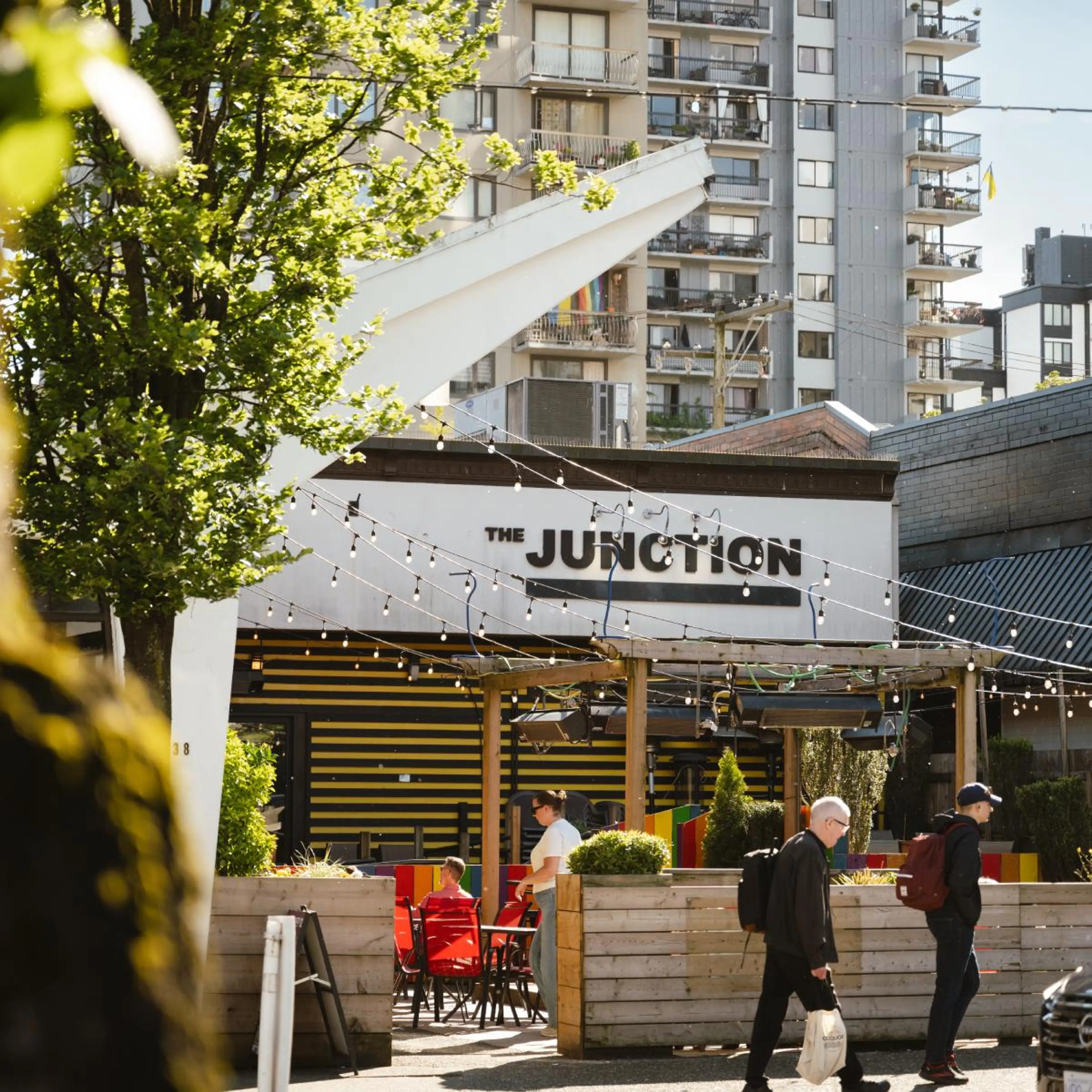 Outdoor seating area of The Junction cafe with string lights, people walking by, and apartment buildings in the background.