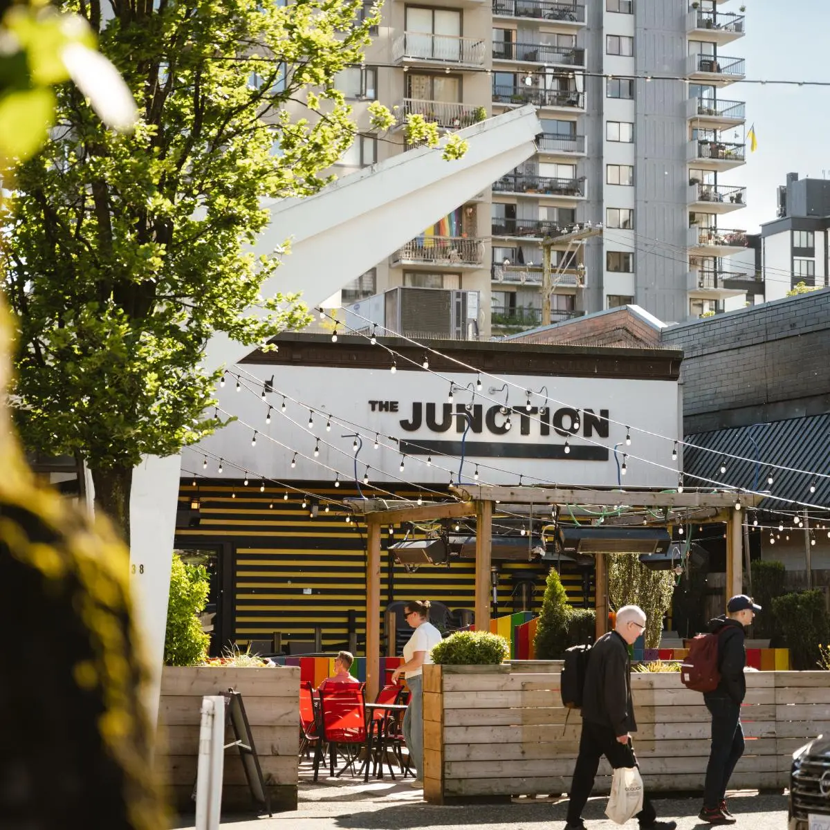 Outdoor seating area of The Junction cafe with string lights, people walking by, and apartment buildings in the background.