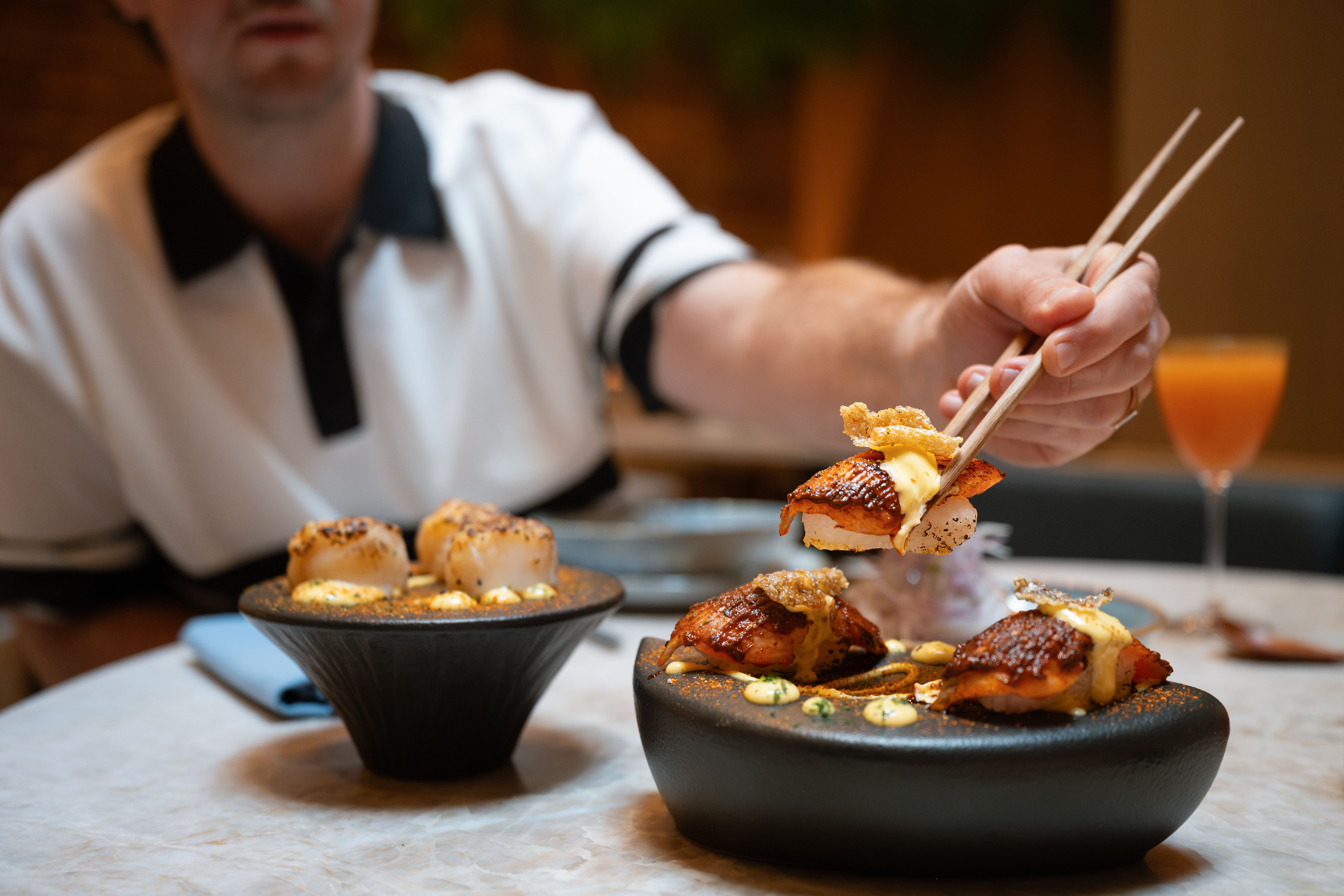 Person using chopsticks to pick up sushi from a black dish on a table with another dish and a drink in the background.