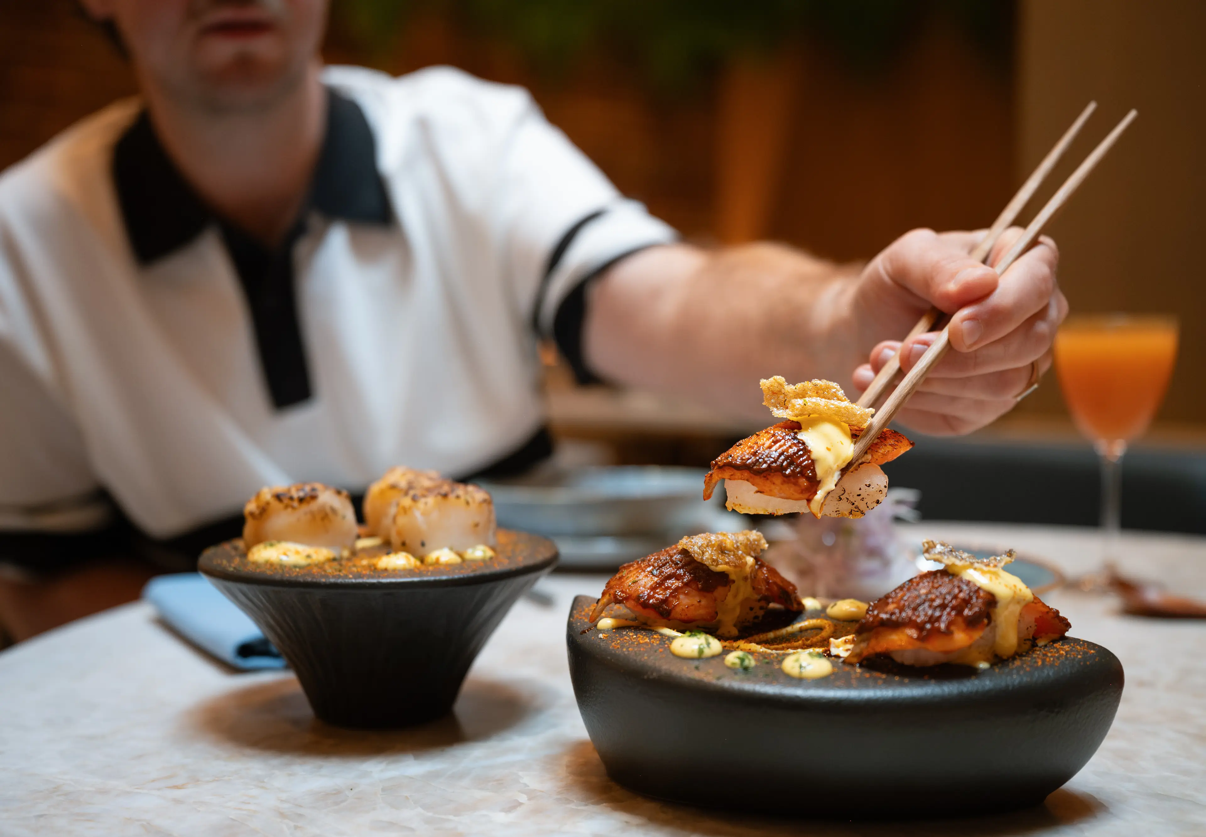 Person using chopsticks to pick up sushi from a black dish on a table with another dish and a drink in the background.