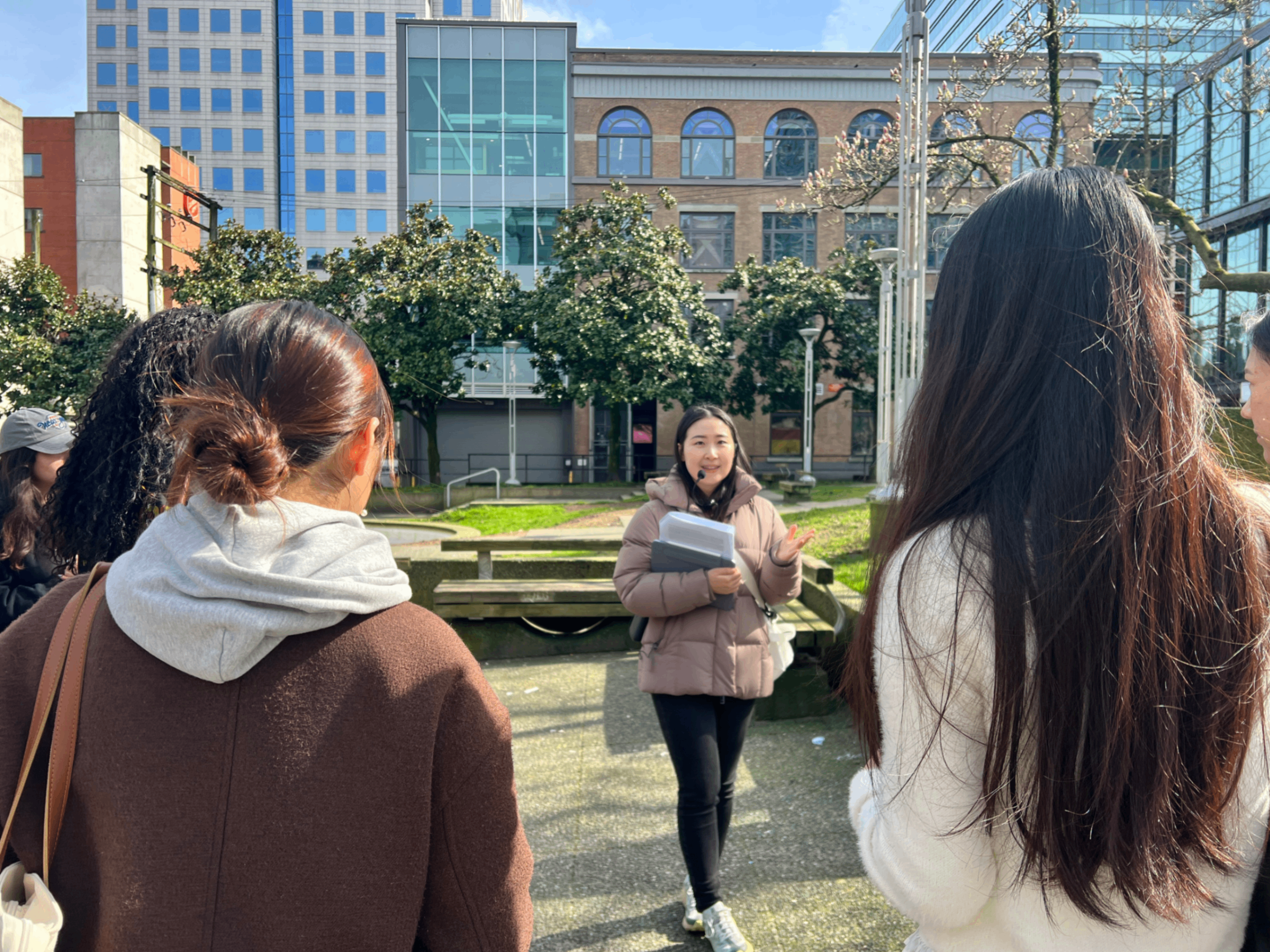 A person in a brown jacket holding papers speaks to a group of five people outdoors in an urban park with buildings and trees in the background.