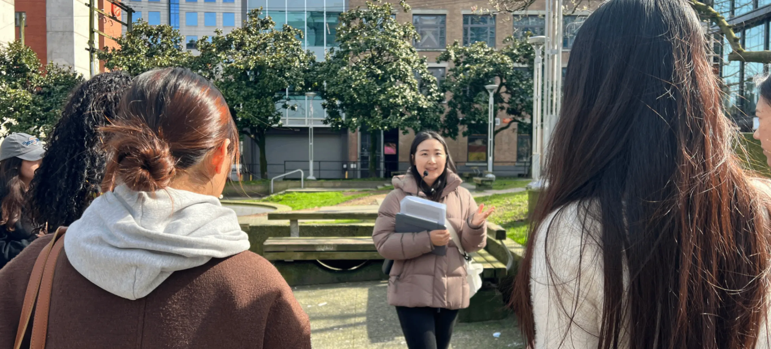 A person in a brown jacket holding papers speaks to a group of five people outdoors in an urban park with buildings and trees in the background.