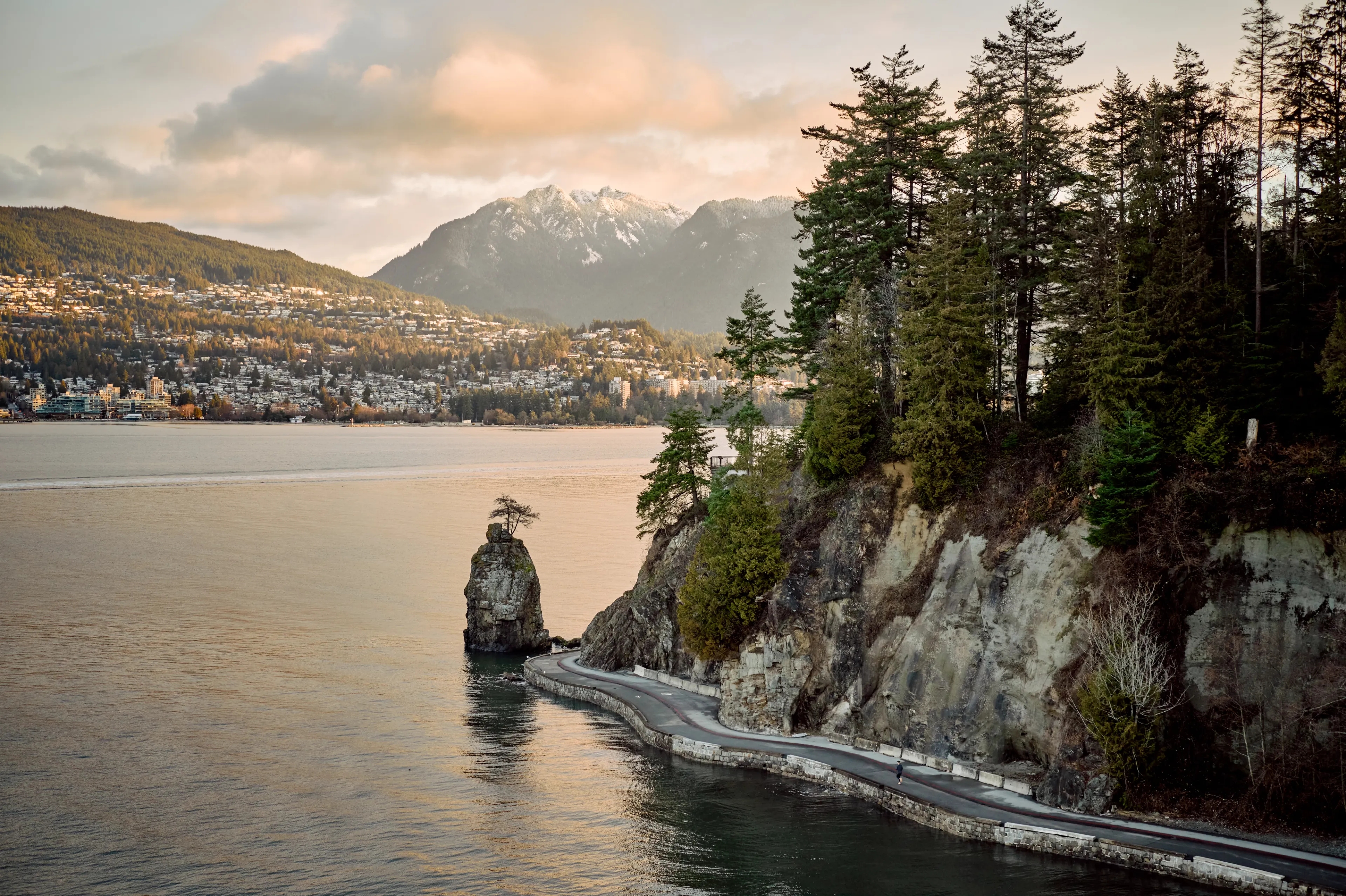 Curved coastal pathway along rocky cliffs with trees, water, and snow-capped mountains in the background at sunset.