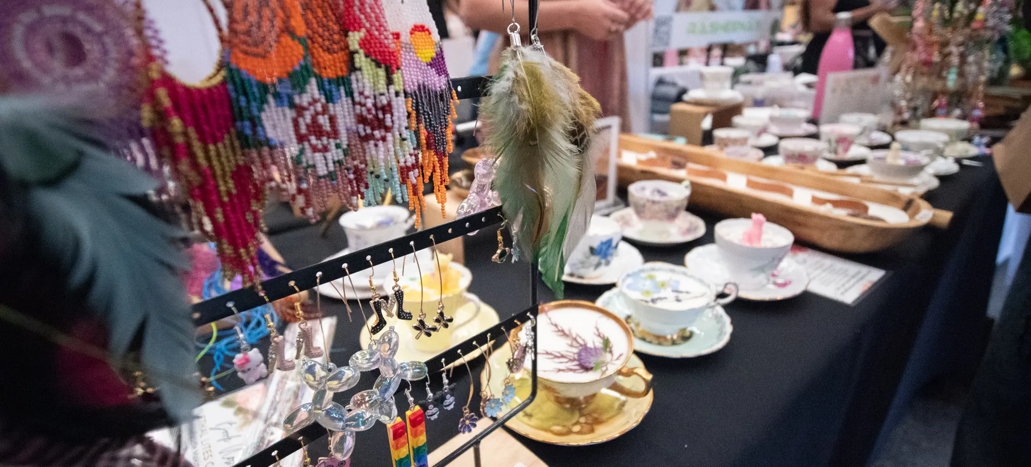 Colorful handmade earrings displayed on a rack with teacups and saucers on a table in the background at a market or craft fair.