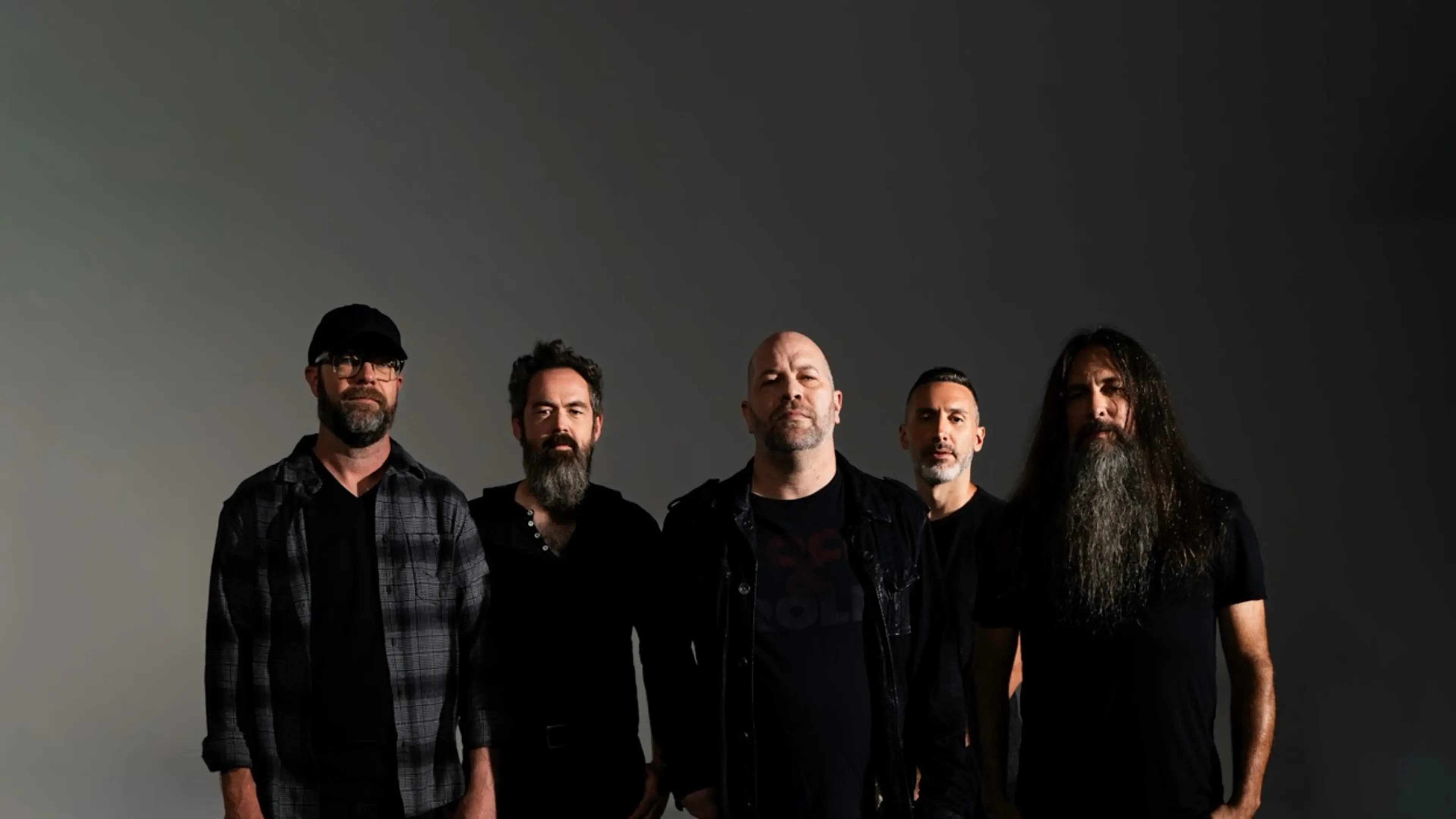 Five people standing against a dark background, wearing dark clothing, with one person wearing a cap and another with a long beard.