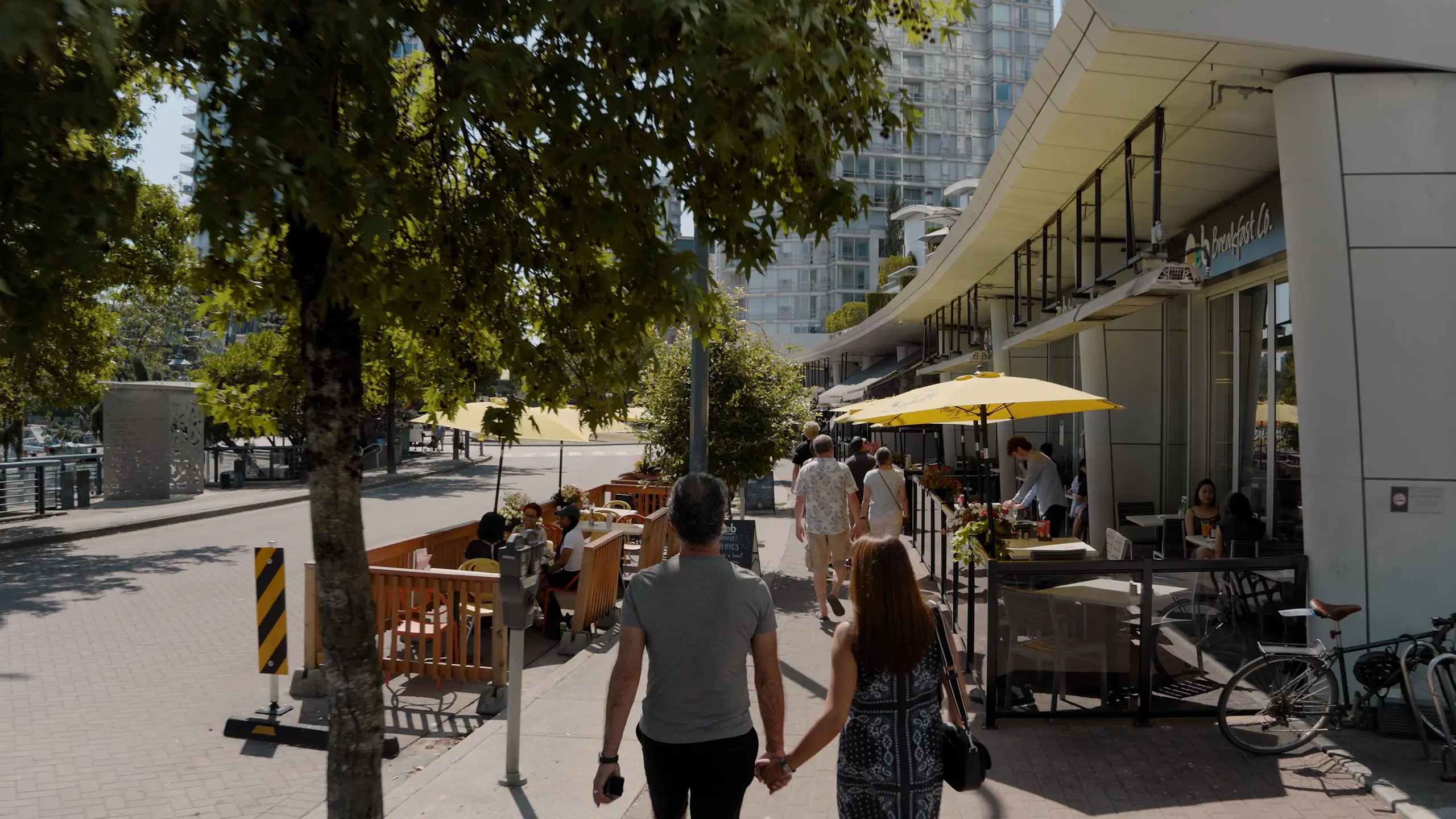 Couple holding hands walking on a sunny city sidewalk past outdoor dining areas with yellow umbrellas and trees.