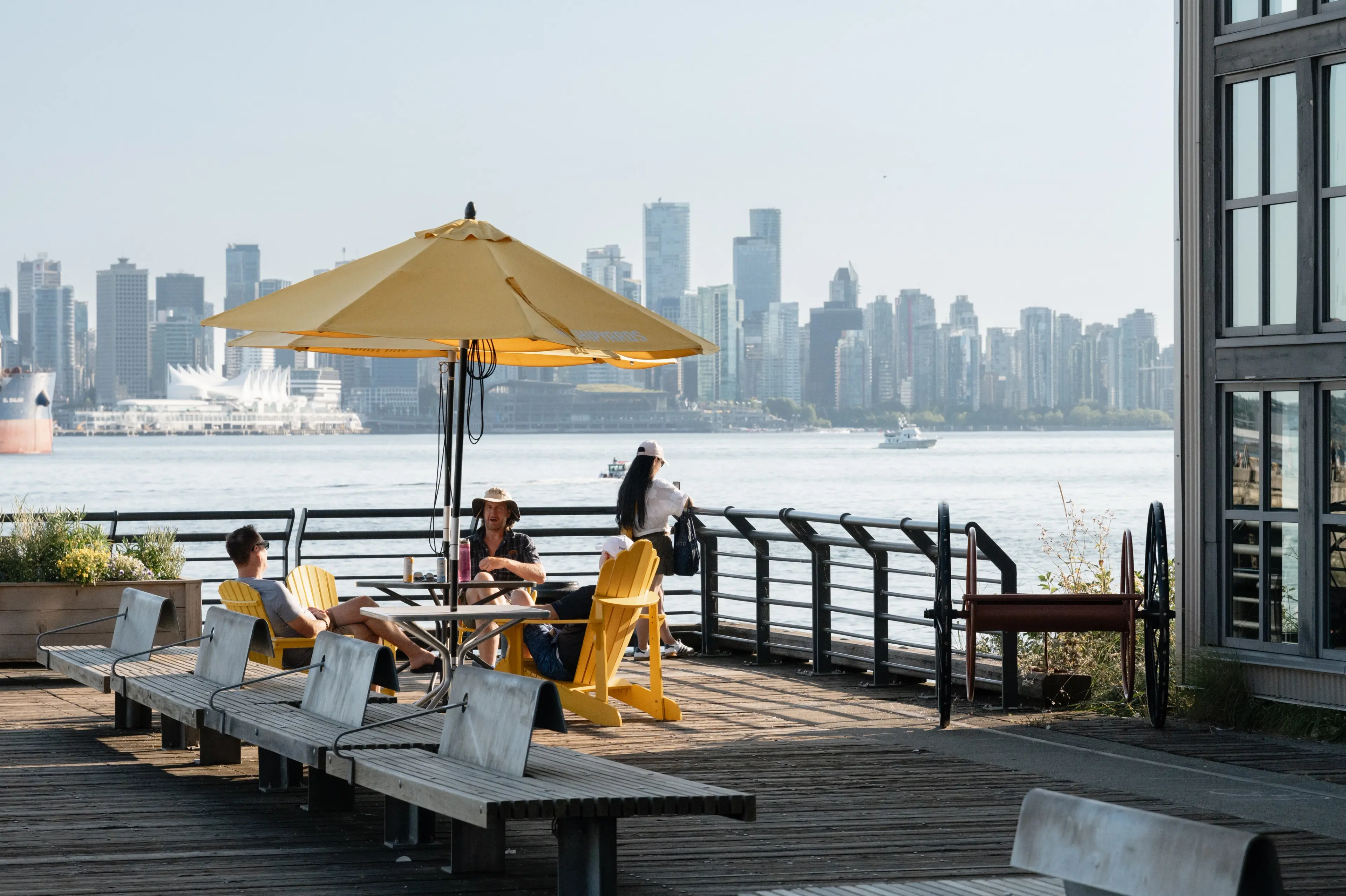 Three people sitting and standing near a yellow umbrella on a wooden pier with a city skyline and water in the background.