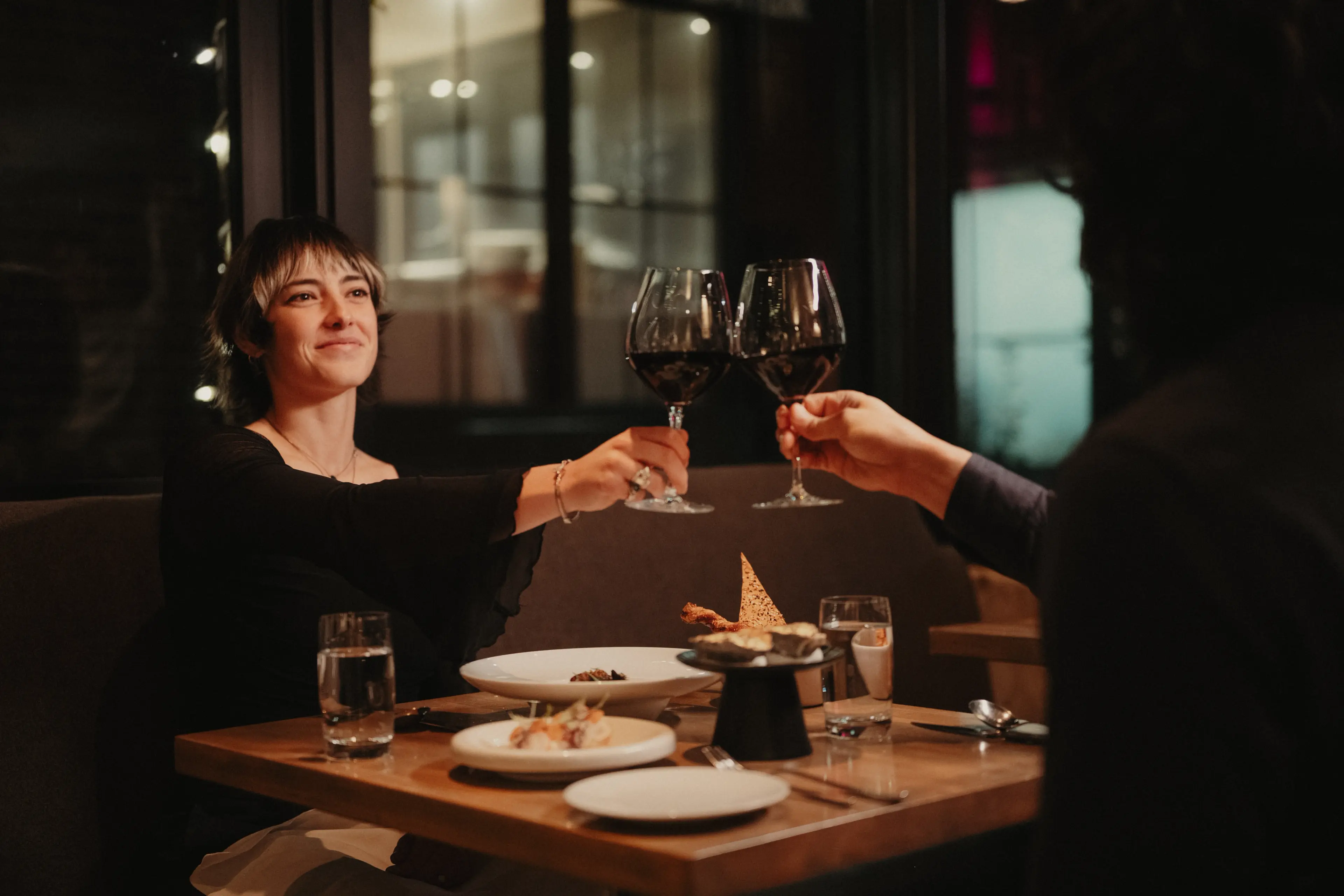 Two people clinking wine glasses over a restaurant table with plates and water glasses at L’Abattoir in Gastown, Vancouver.