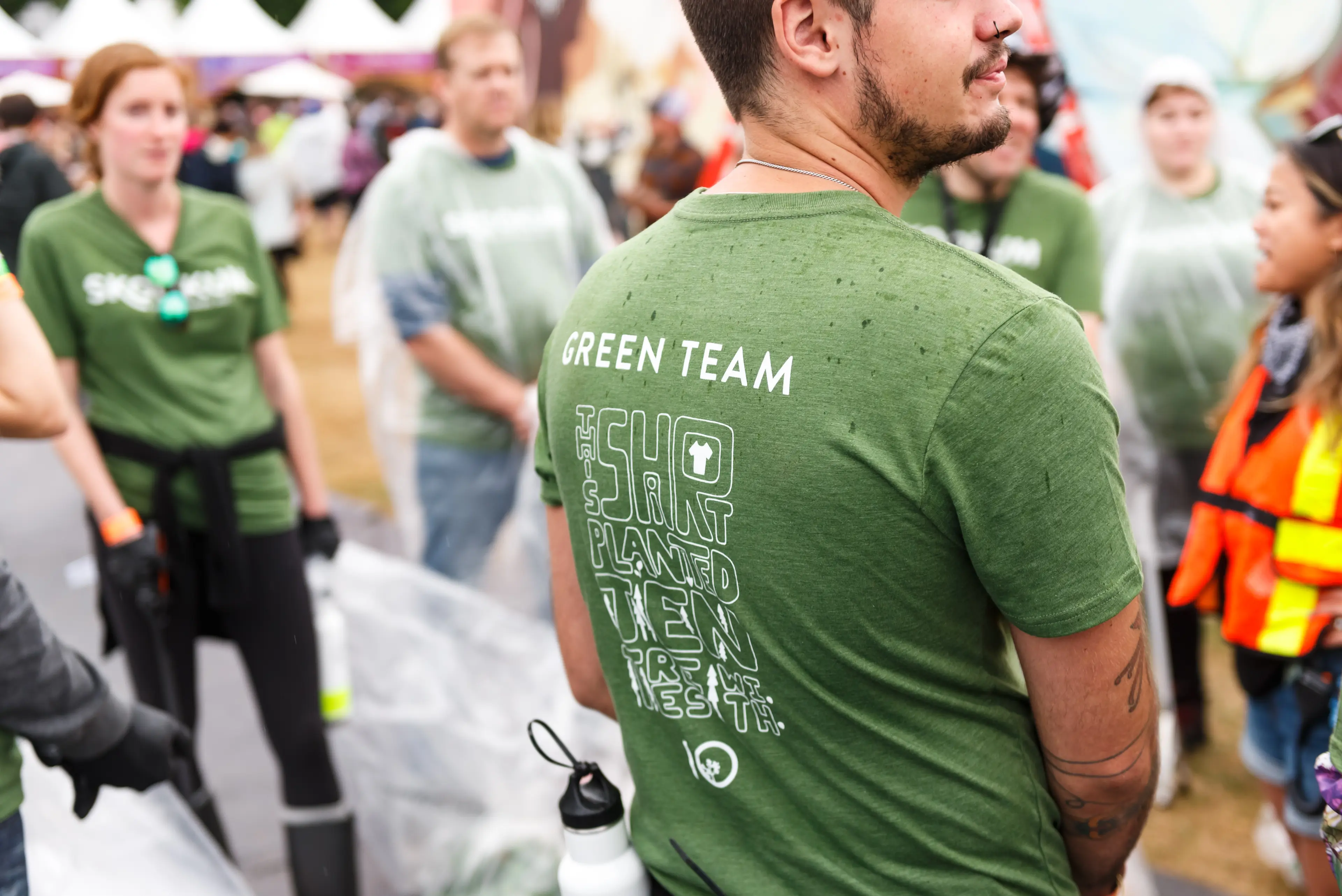 The back of a SKOOKUM Music Festival volunteer, showing their Green Team t-shirt. Other volunteers are around them.