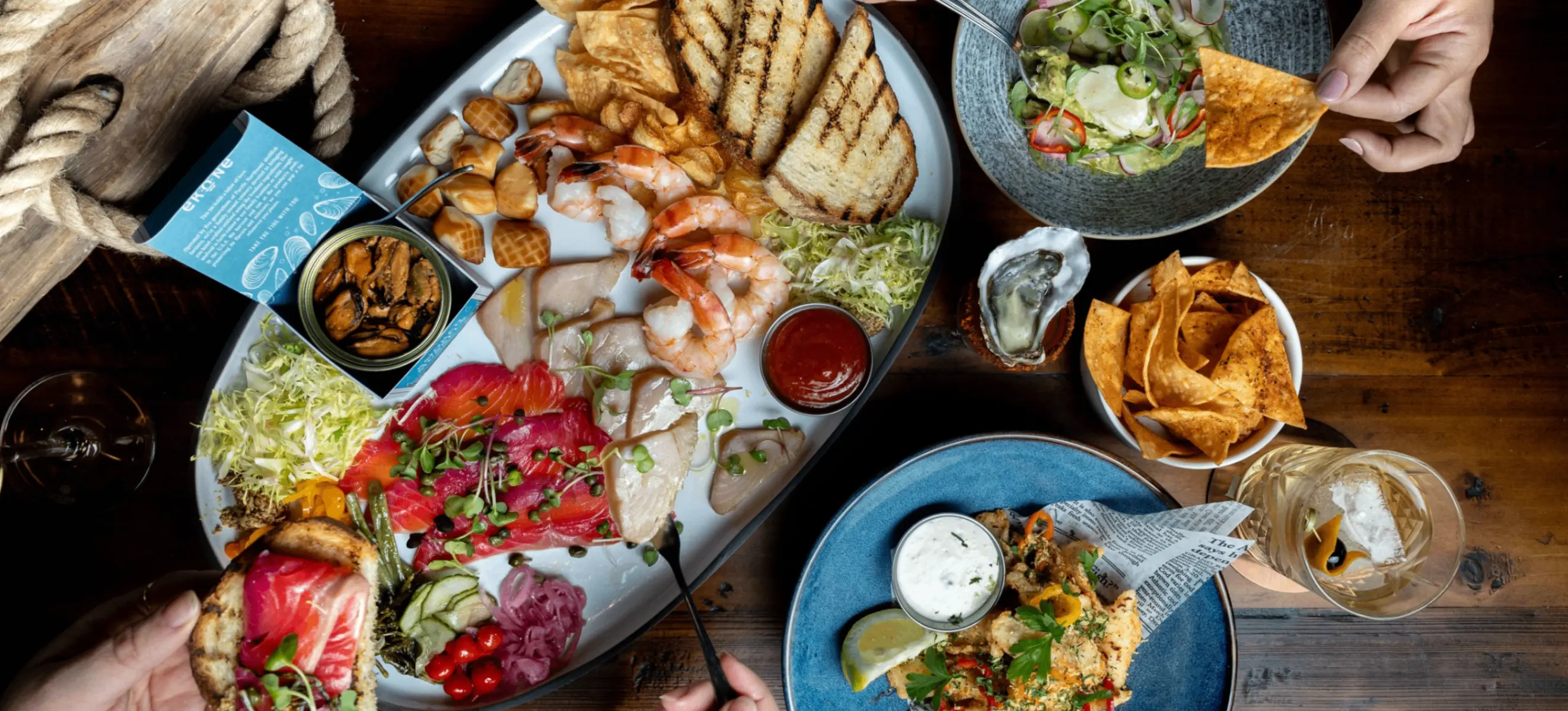 Overhead view of a seafood platter with shrimp, smoked fish, grilled bread, and sauces, with hands holding a chip and a piece of bread with toppings.