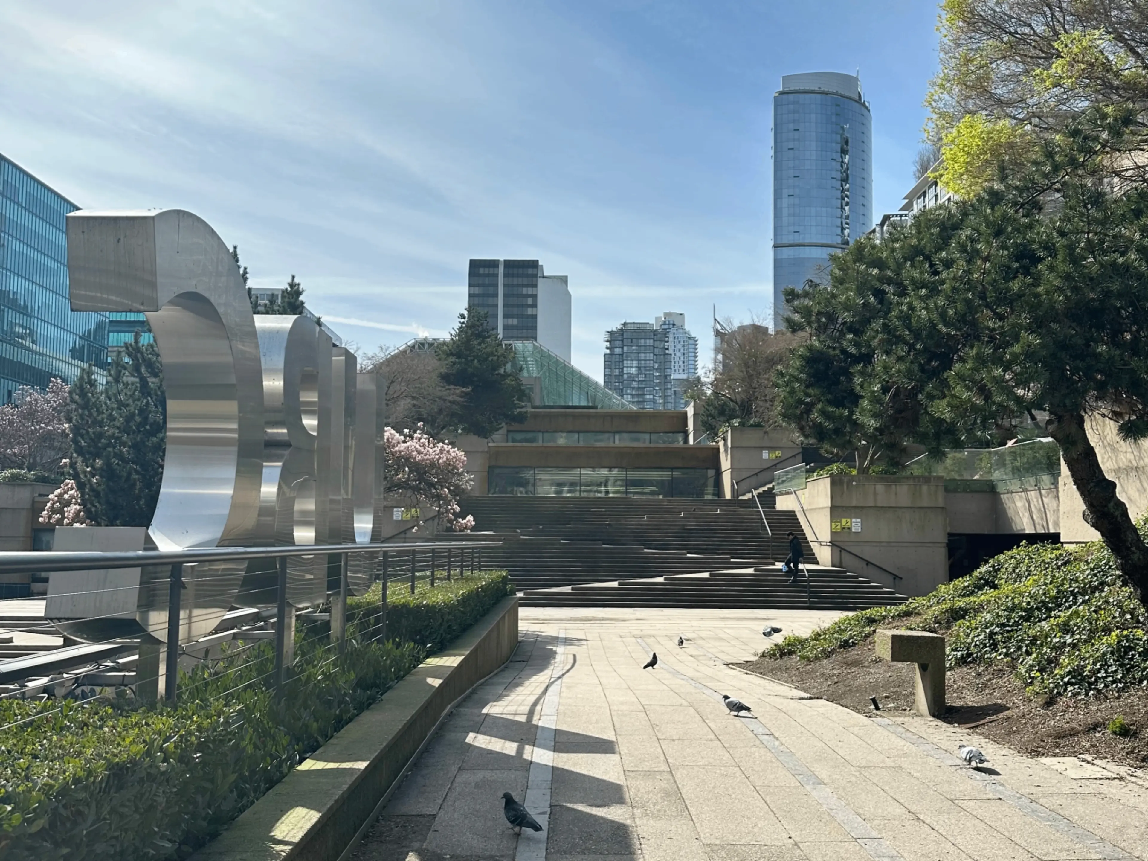 Outdoor urban scene with large metallic sculpture, stairs, trees, pigeons on pathway, and tall buildings in the background.