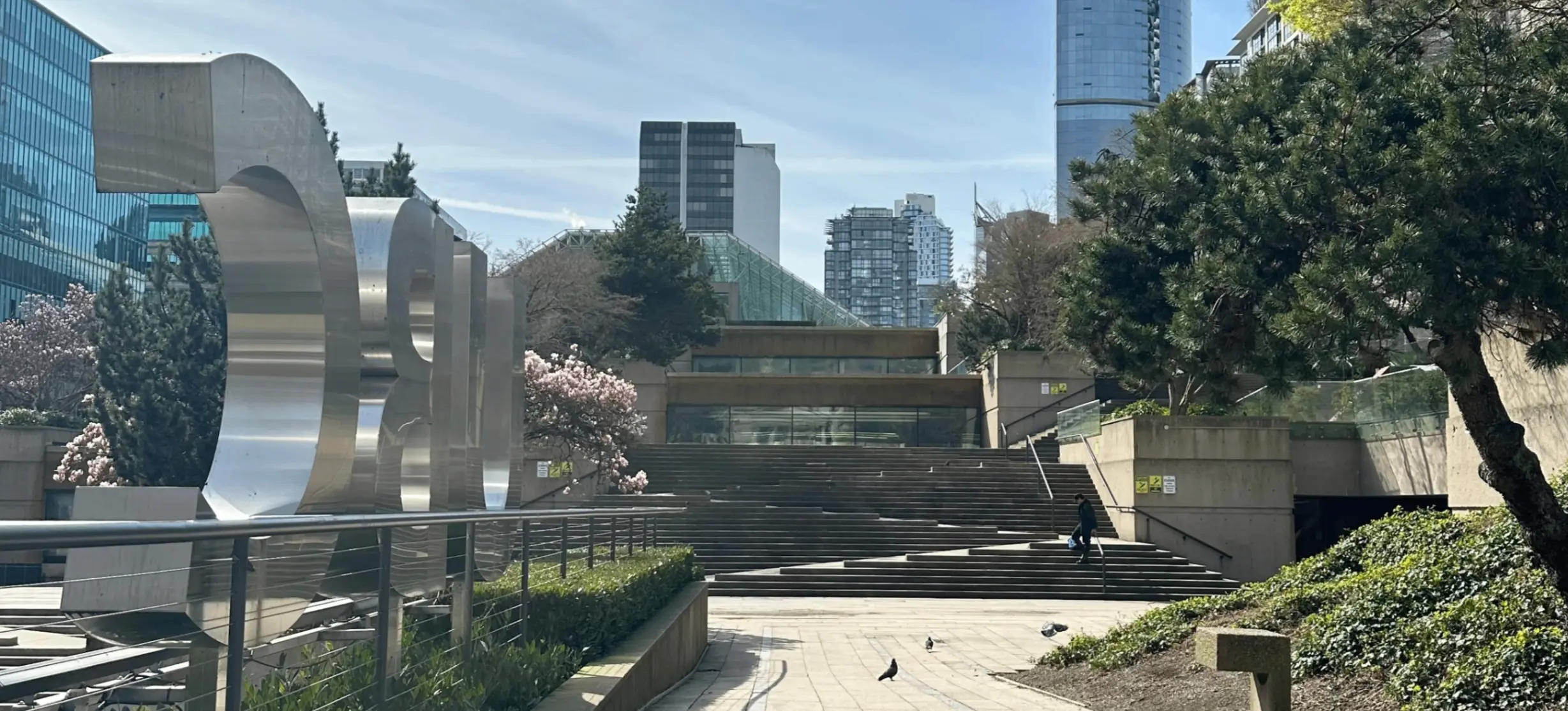 Outdoor urban scene with large metallic sculpture, stairs, trees, pigeons on pathway, and tall buildings in the background.