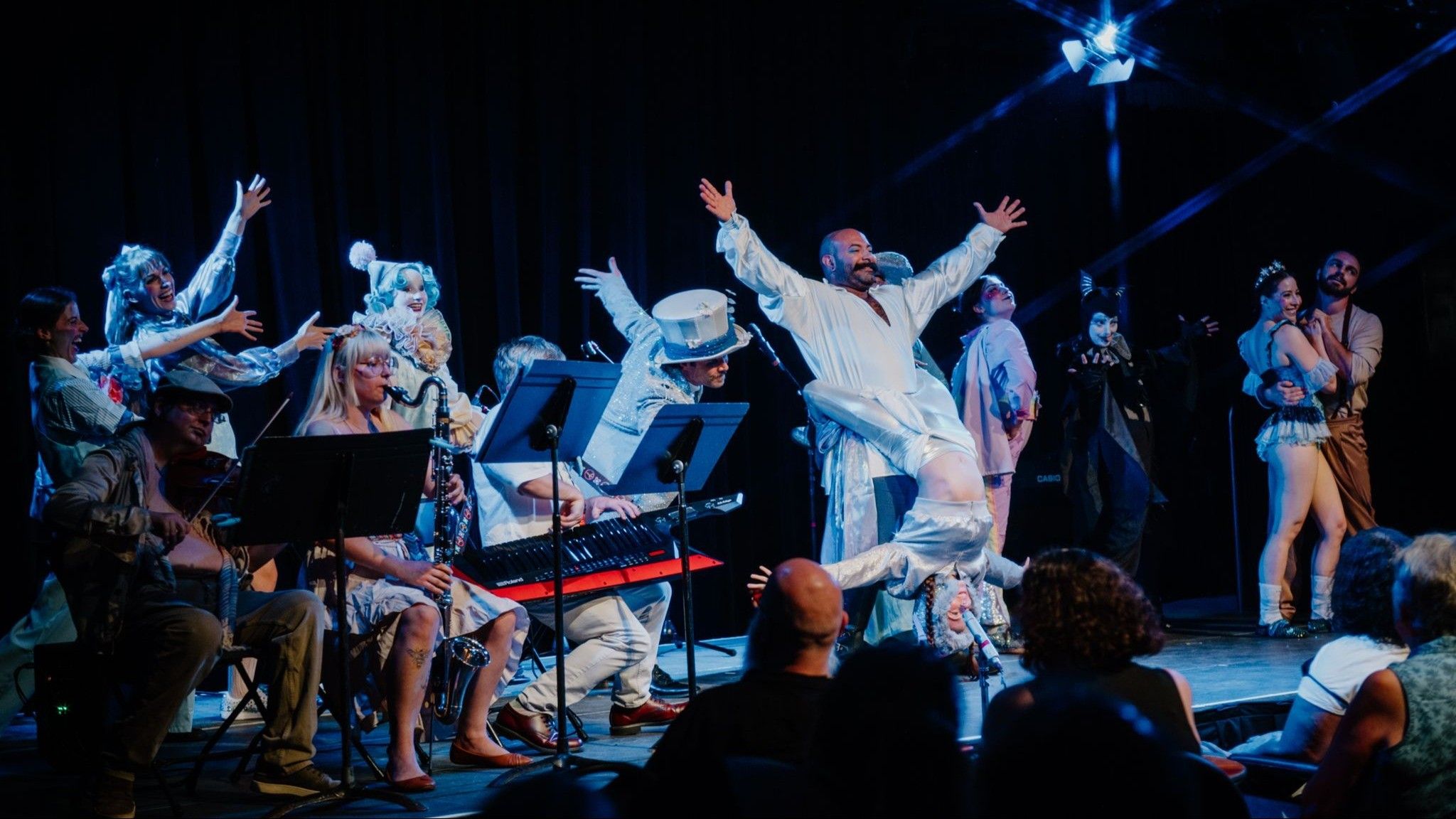 A theatrical performance with musicians playing instruments and actors in colorful costumes on stage, with an audience watching.
