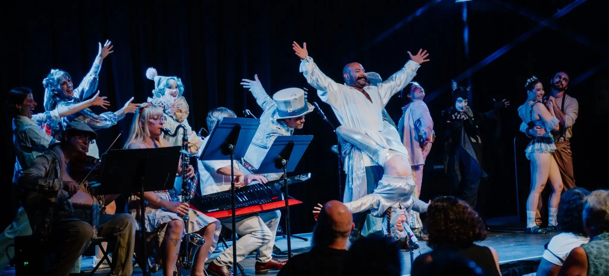 A theatrical performance with musicians playing instruments and actors in colorful costumes on stage, with an audience watching.