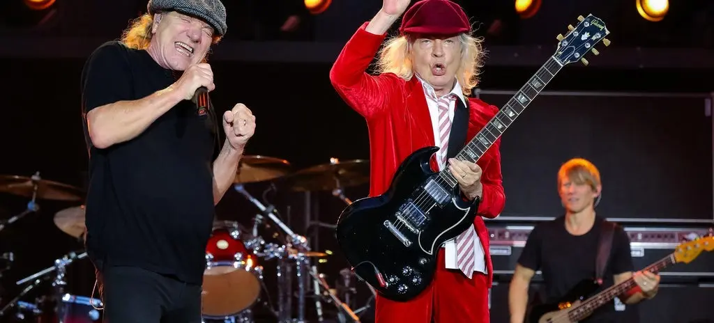 Three musicians performing on stage with one singing into a microphone, one playing guitar in a red suit and hat, and one playing bass guitar.