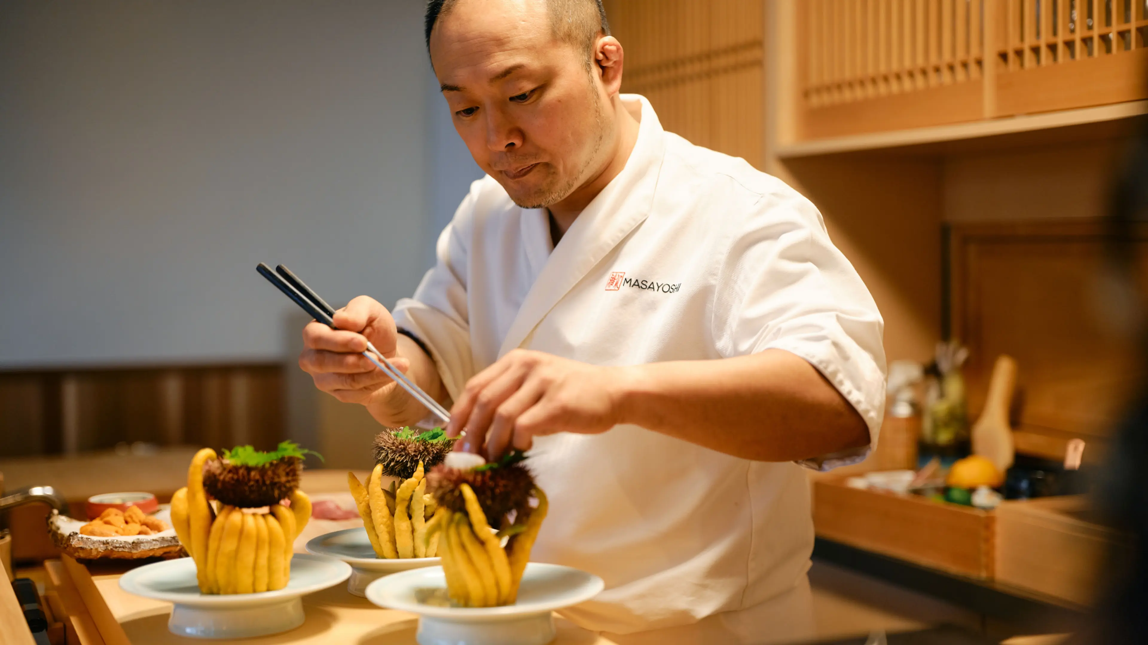 A chef in a white uniform carefully preparing dishes with sea urchins and yellow garnishes on white plates in a kitchen.