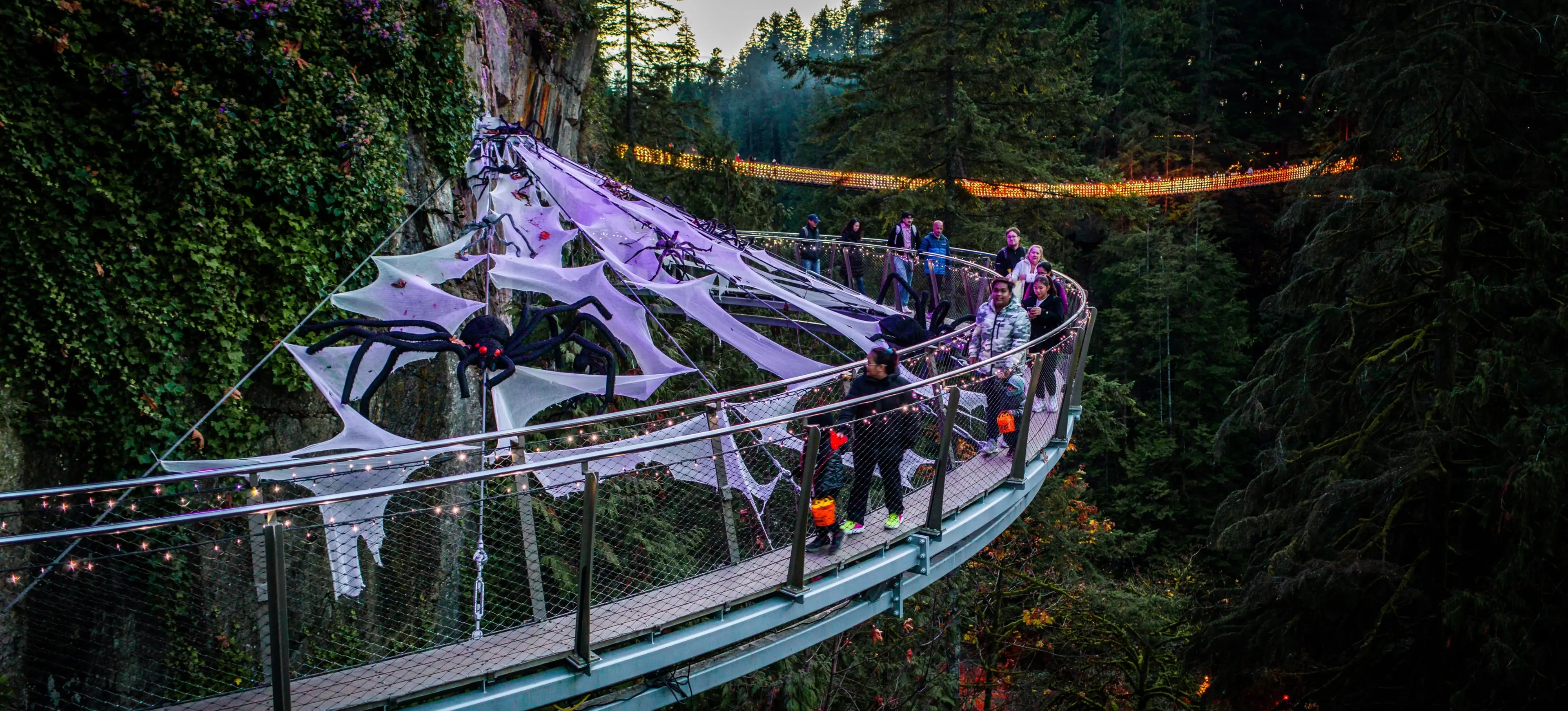 People walking on a curved suspension bridge decorated with large fake spider webs and spiders, surrounded by forest at dusk.