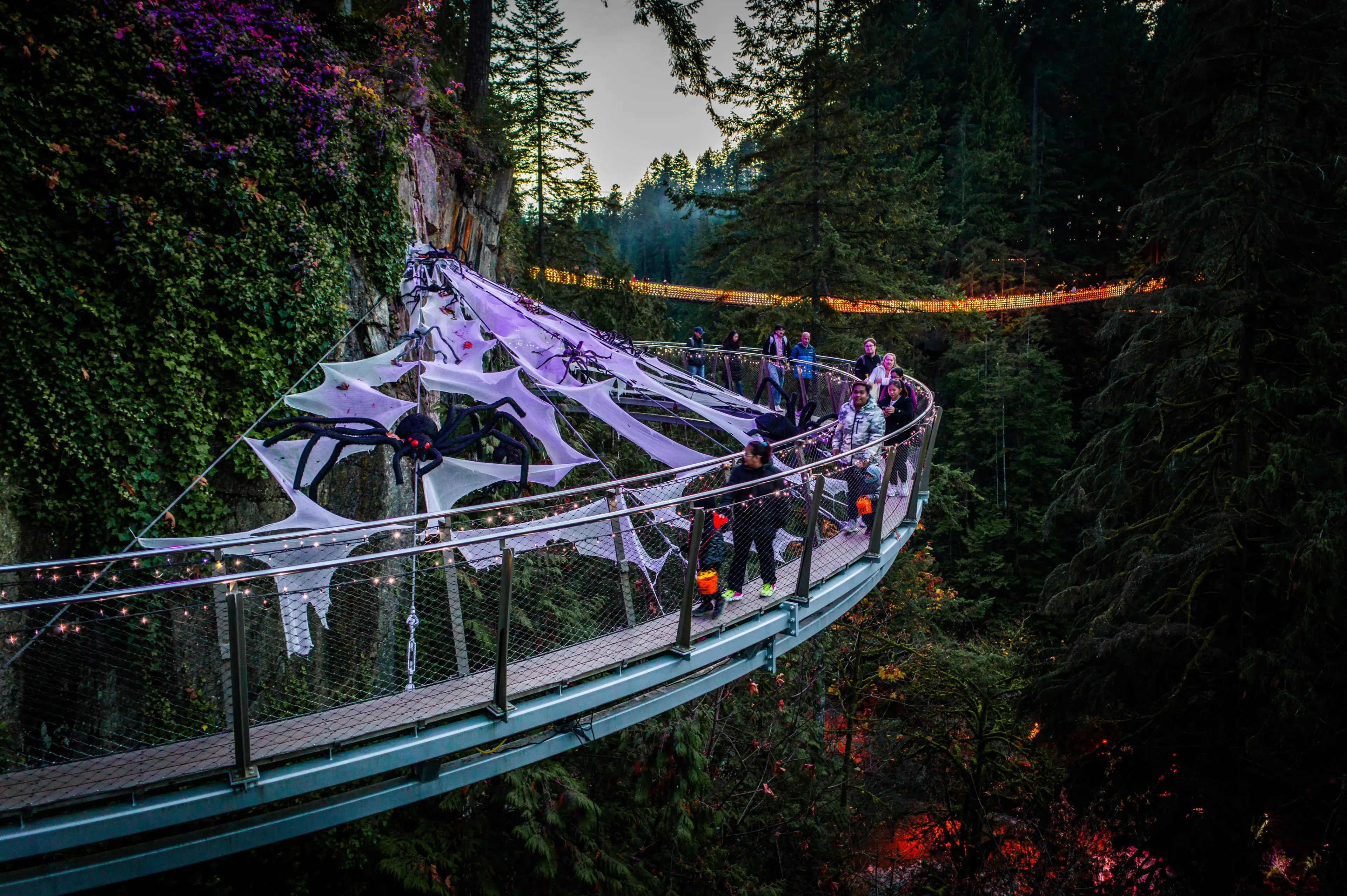People walking on a curved suspension bridge decorated with large fake spider webs and spiders, surrounded by forest at dusk.