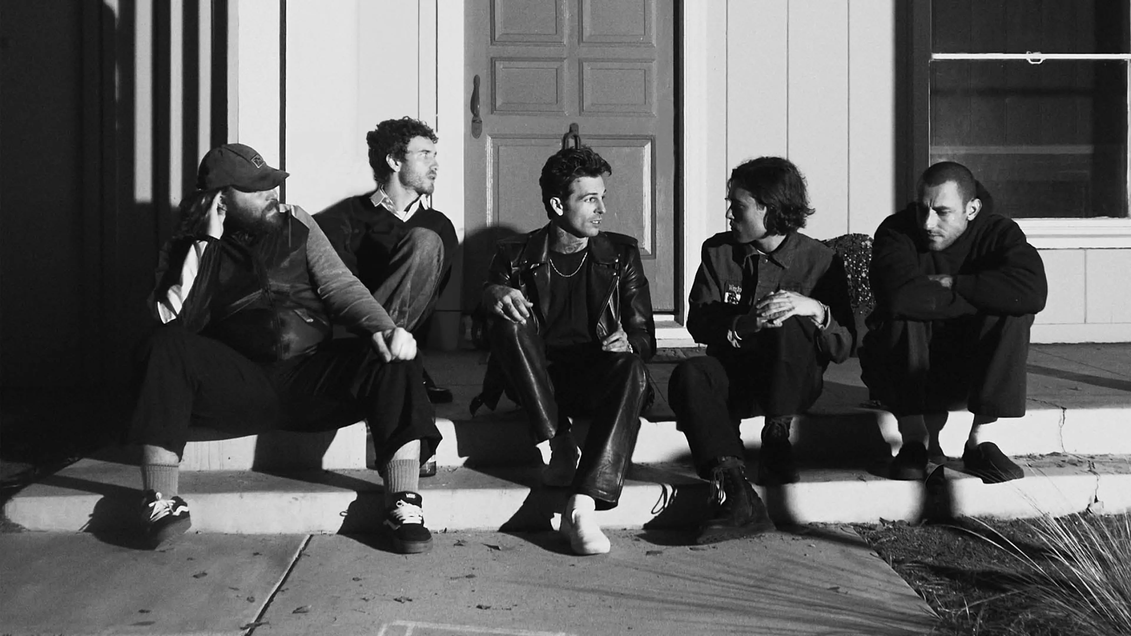 Five people sitting on outdoor steps in front of a door, engaged in conversation, in black and white.