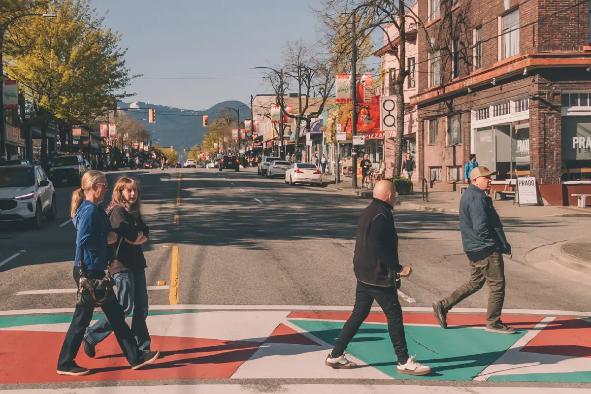 Two people walking on a colorful crosswalk from left to right, and two people walking from right to left on a sunny street with shops and cars.