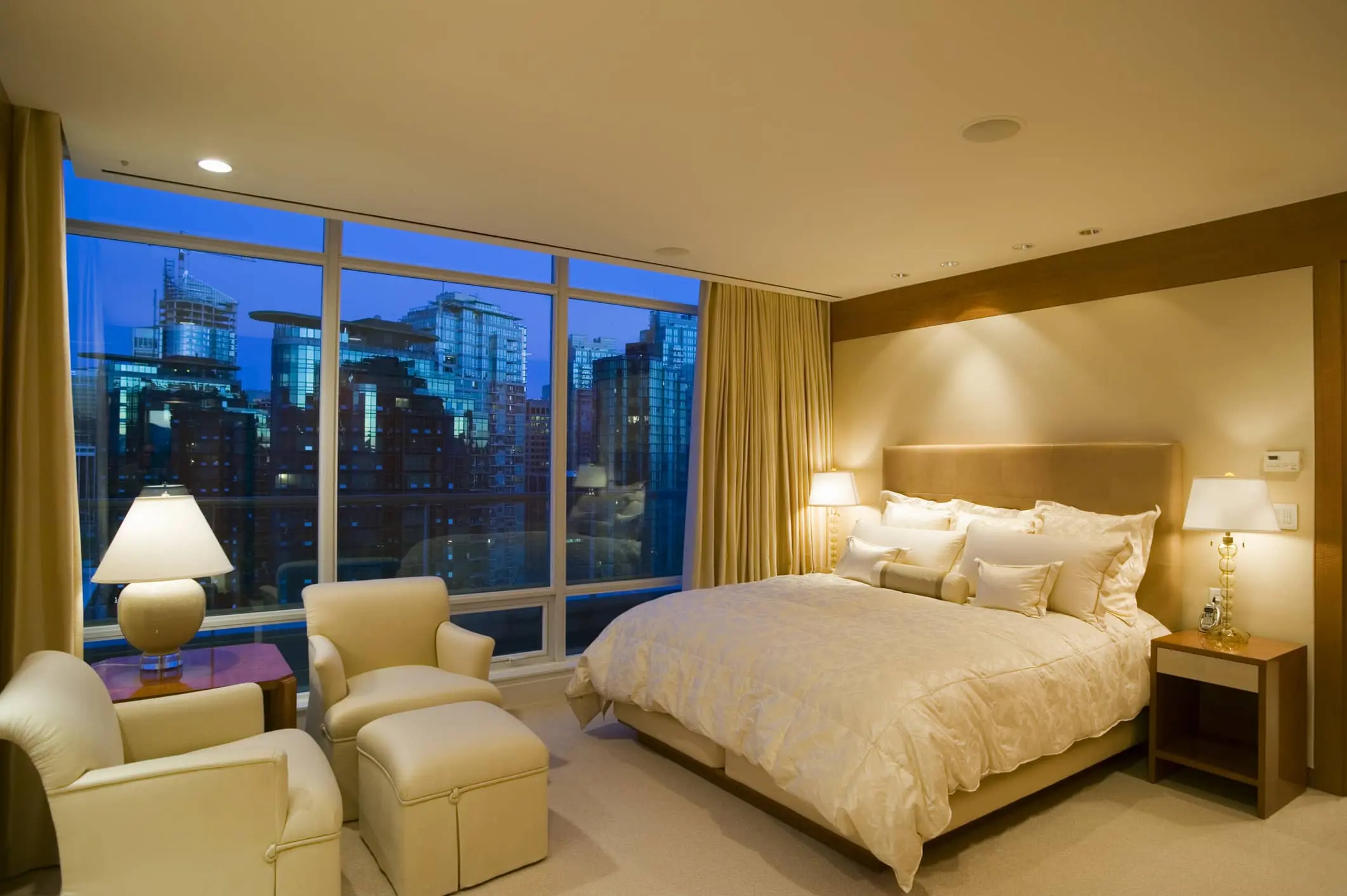 A hotel room with a large bed and two leather chairs looks out on the city.