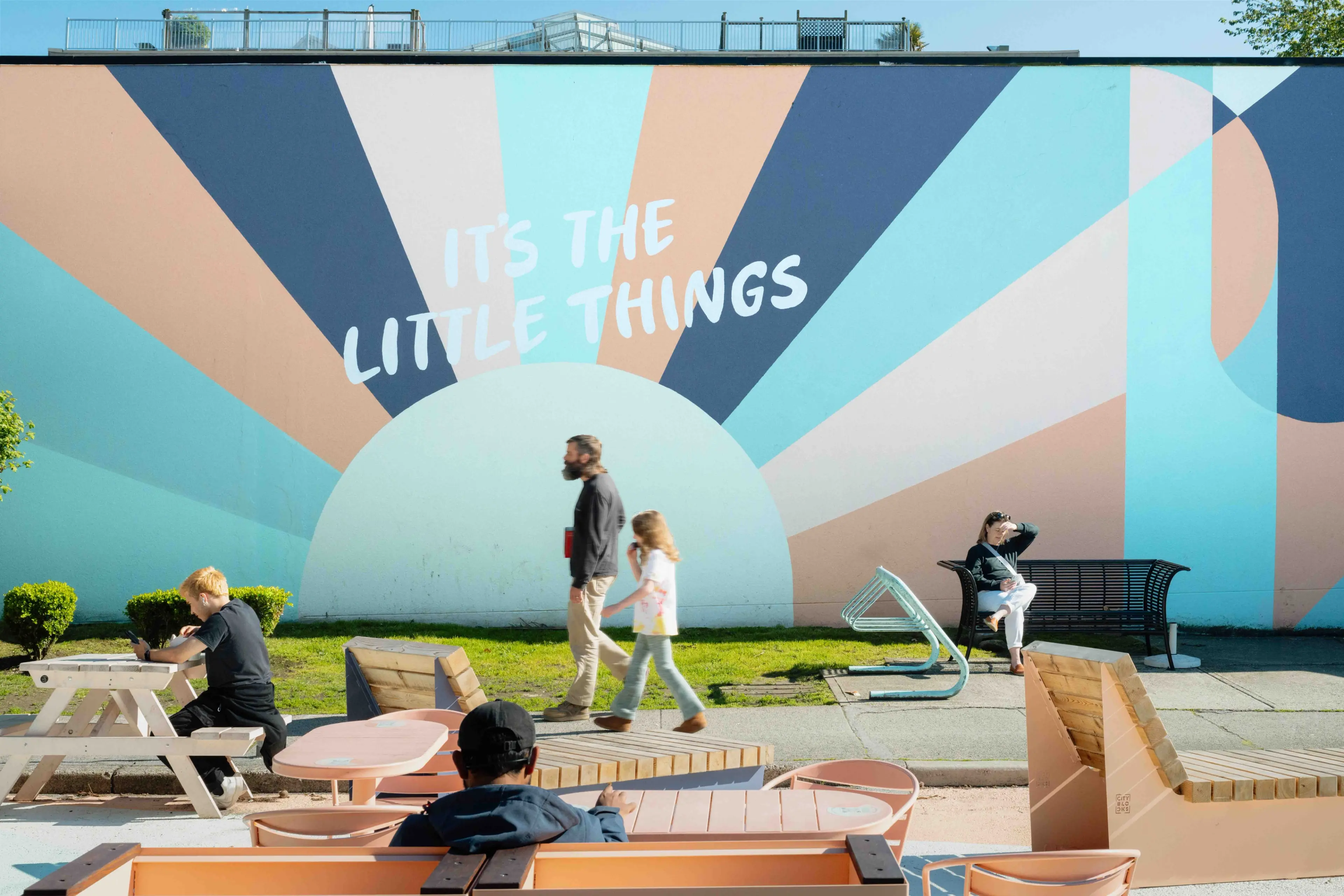 A colorful mural with sunburst patterns and the words ITS THE LITTLE THINGS painted on a wall in Vancouver City. People walk and sit in front of the mural on a sunny day, enjoying outdoor seating visible in the foreground.
