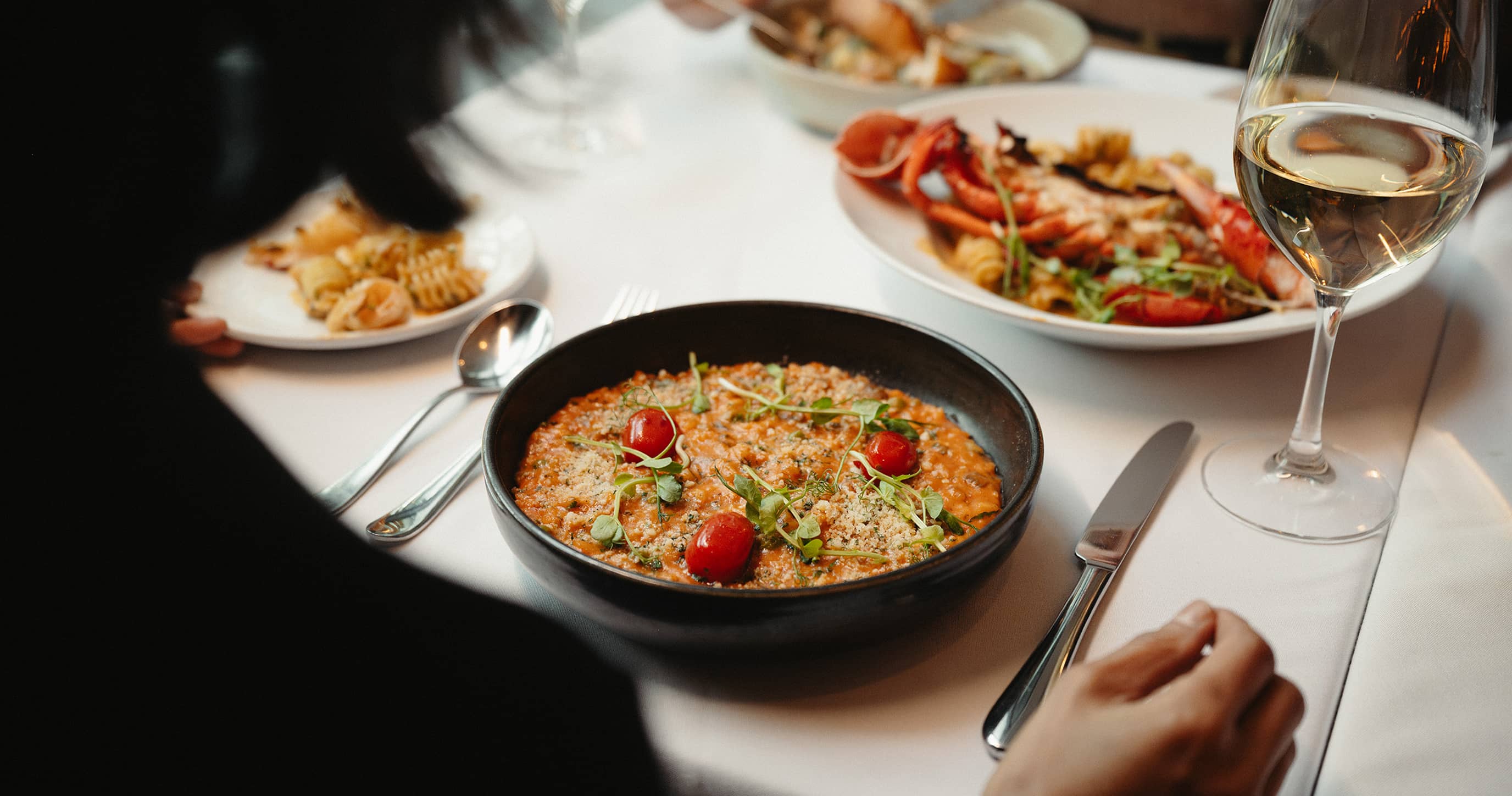 Person sitting at a table with a bowl of risotto garnished with cherry tomatoes and herbs, a glass of white wine, and plates of seafood pasta.