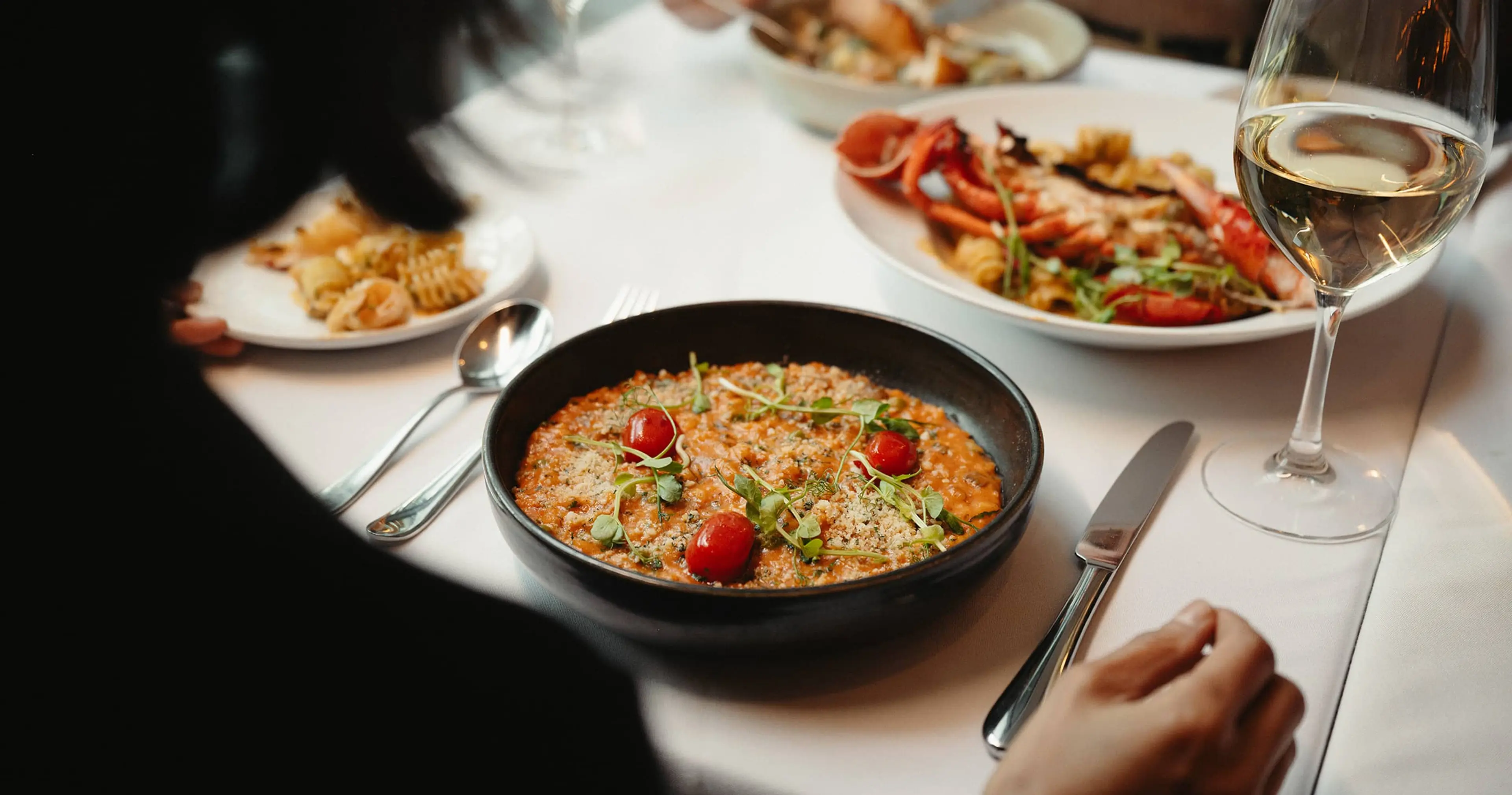 Person sitting at a table with a bowl of risotto garnished with cherry tomatoes and herbs, a glass of white wine, and plates of seafood pasta.