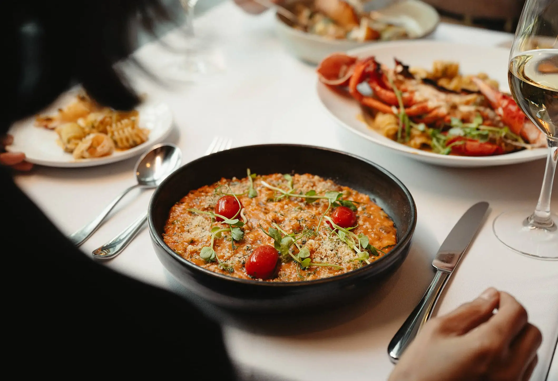 Person sitting at a table with a bowl of risotto garnished with cherry tomatoes and herbs, a glass of white wine, and plates of seafood pasta.
